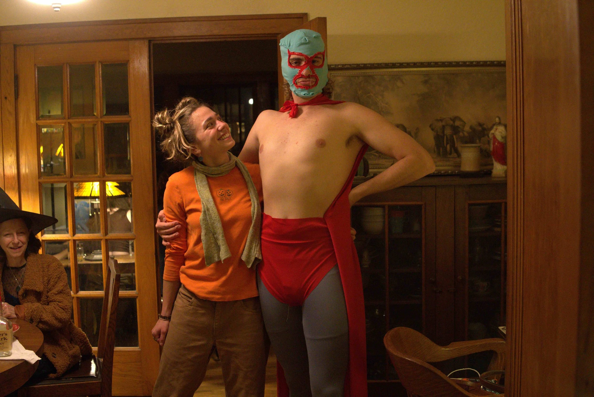 A person wearing a blue luchador mask and a red cape stands next to a smiling woman in an orange dress inside a room with wooden walls and glass-paneled doors.