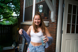 A young woman smiling outside near a brick house, holding a glass of water in one hand and a jacket in the other.