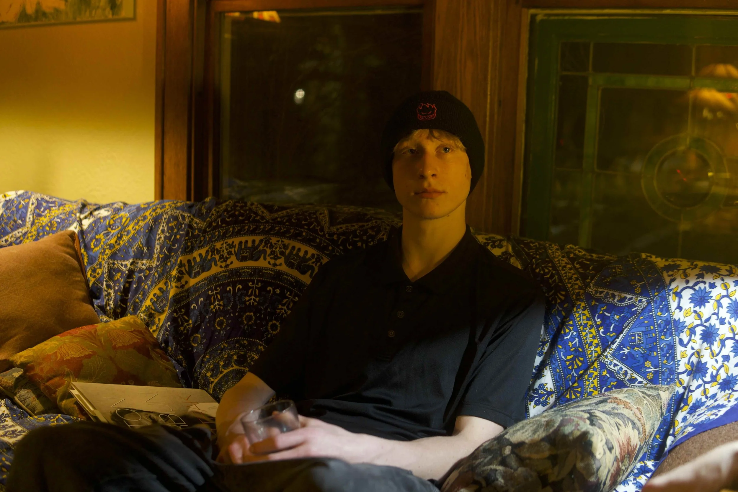 A young man with light hair and a black beanie sits on a colorful patterned couch in a cozy living room holding a glass, with a window behind him showing a night scene.