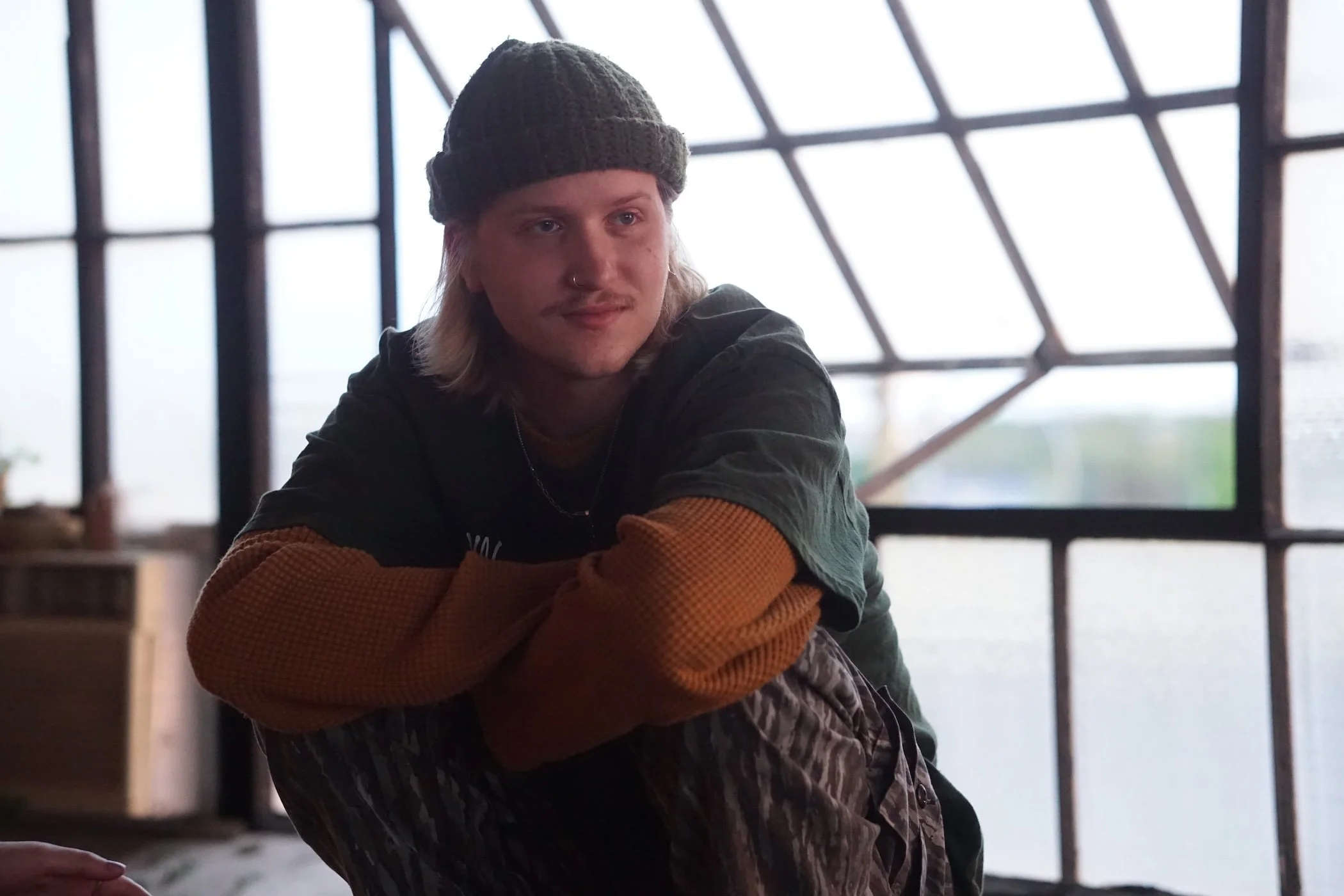 A person with long hair, wearing a dark beanie and a nose ring, is hugging someone in an indoor setting with large window panes in the background.