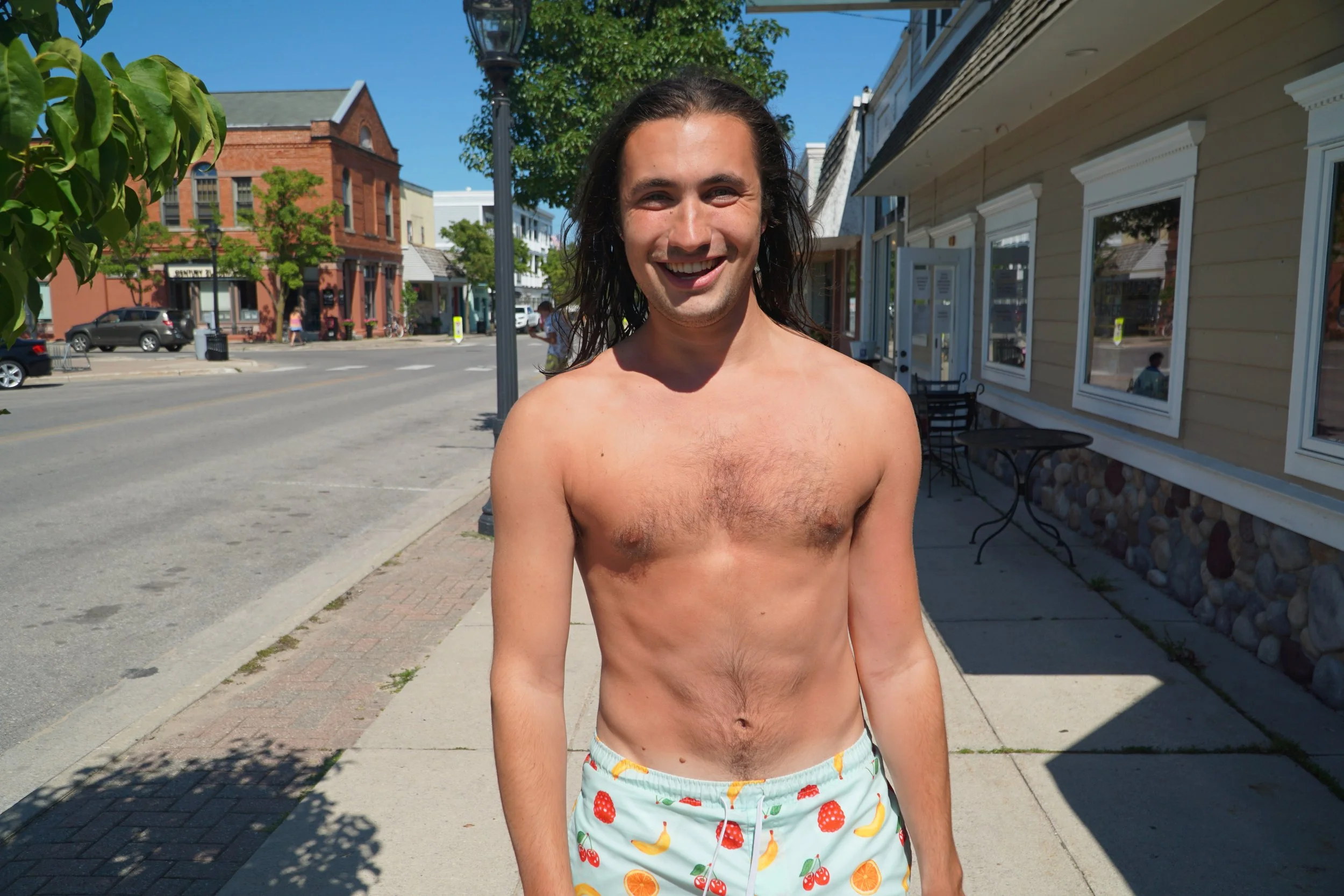 A shirtless man with long dark hair smiling on a sidewalk in front of shops and cars on a sunny day.