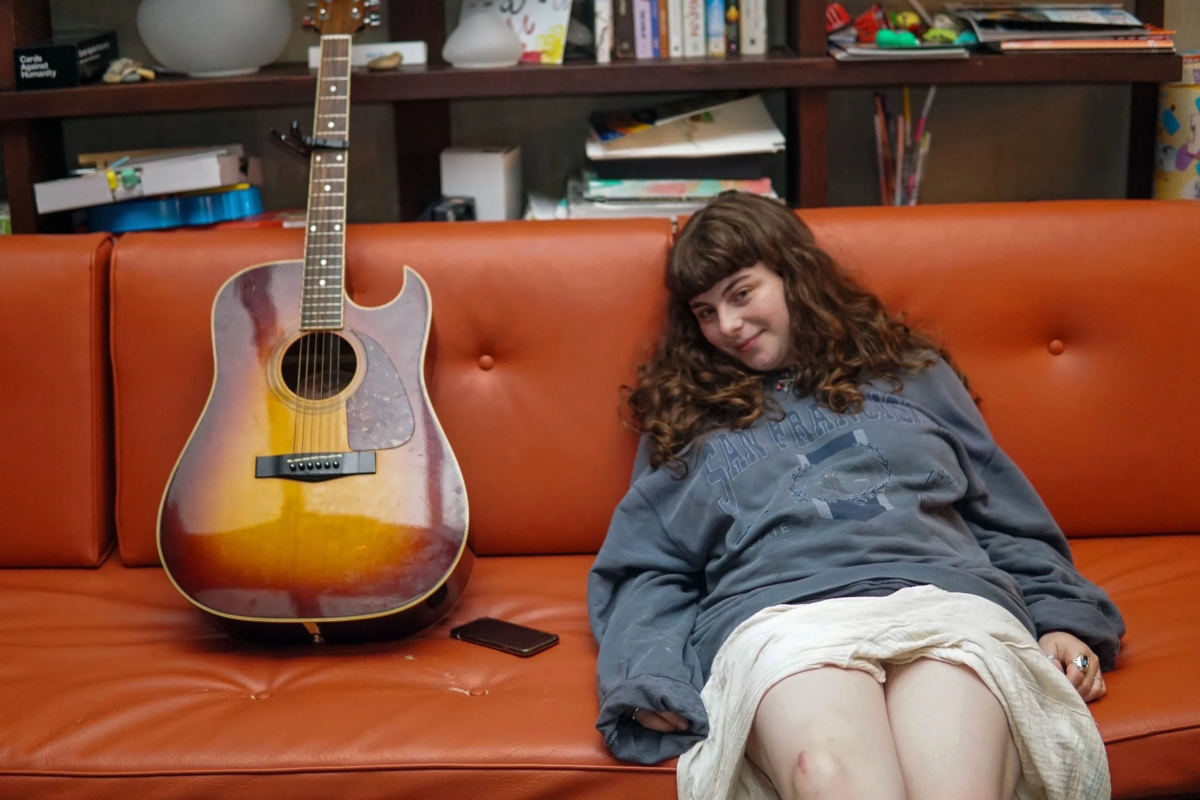 A young girl with curly brown hair, wearing a gray sweatshirt and beige shorts, sitting on an orange leather couch with her head tilted slightly to the side and smiling. An acoustic guitar with a sunburst finish rests against the back of the couch, next to her. In the background, a wooden bookshelf filled with books, papers, and various objects.
