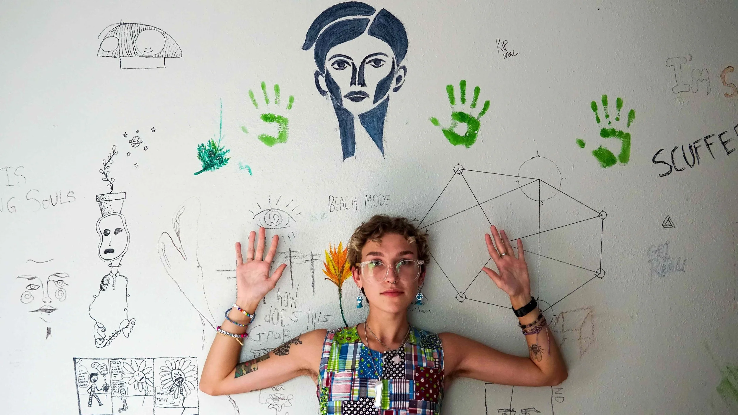 A person with short curly hair, glasses, and colorful clothing standing in front of a white wall covered with various drawings, doodles, and writings, holding up both hands with open palms facing the camera.