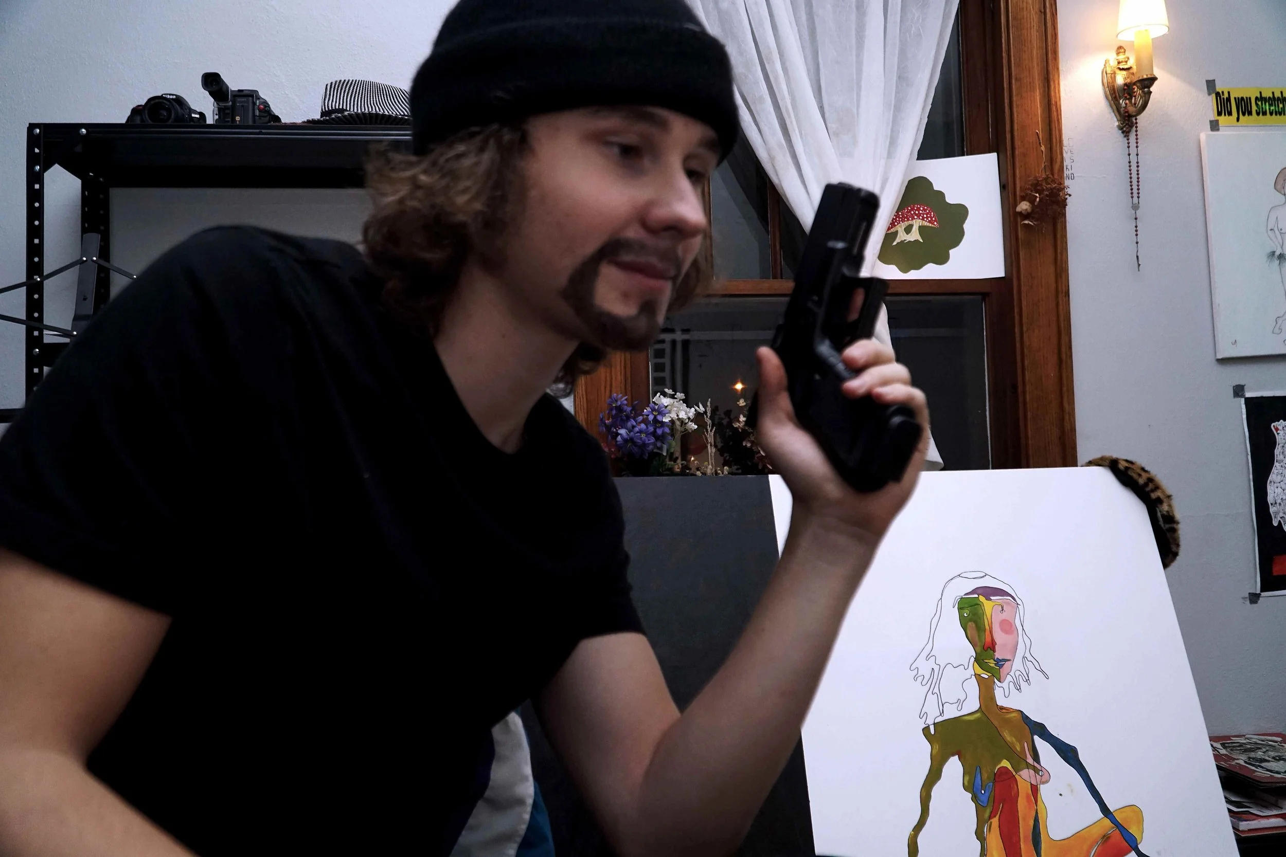 Young man with long hair, beard, and mustache wearing a black beanie looking at his phone, in a room with artwork and a shelf with cameras in the background.