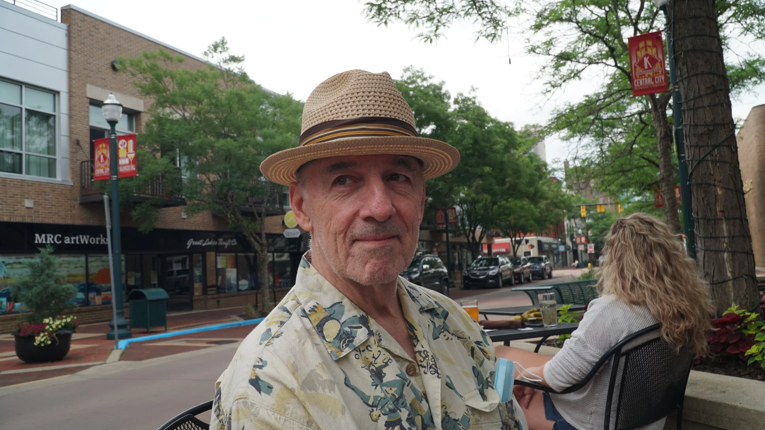A man, Tamara Skidmore's husband George Kuttner, wearing a straw hat and floral shirt sitting outdoors at a table with a woman with curly blonde hair, in an urban area with trees, storefronts, parked cars, and street signs in the background.