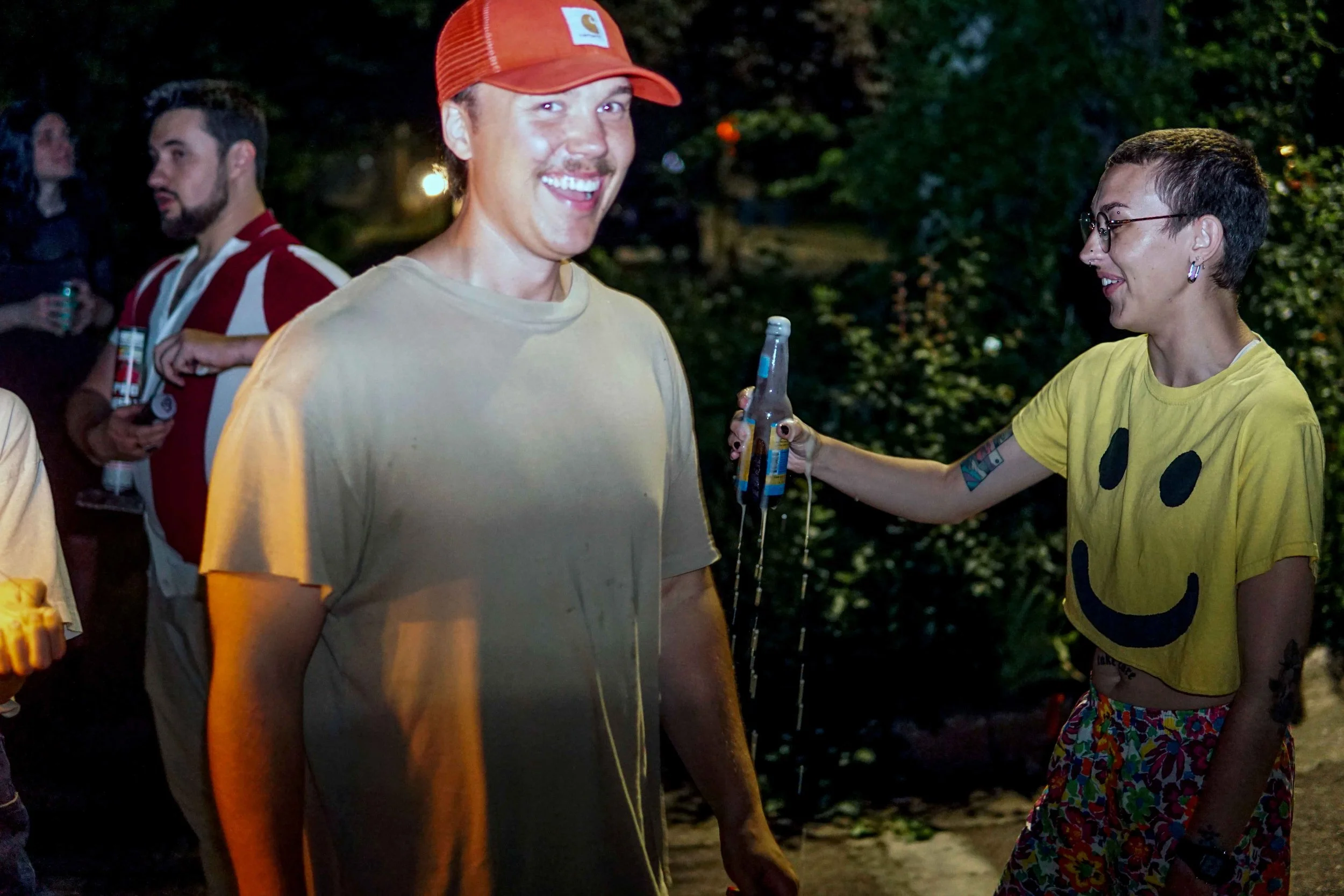 A group of people at an outdoor gathering at night, smiling and holding drinks. One man in the foreground is wearing a beige shirt and a red cap. A woman to his right is wearing a yellow shirt with a smiley face design and colorful shorts, with tattoos on her arm. The background shows trees and blurred lighting.