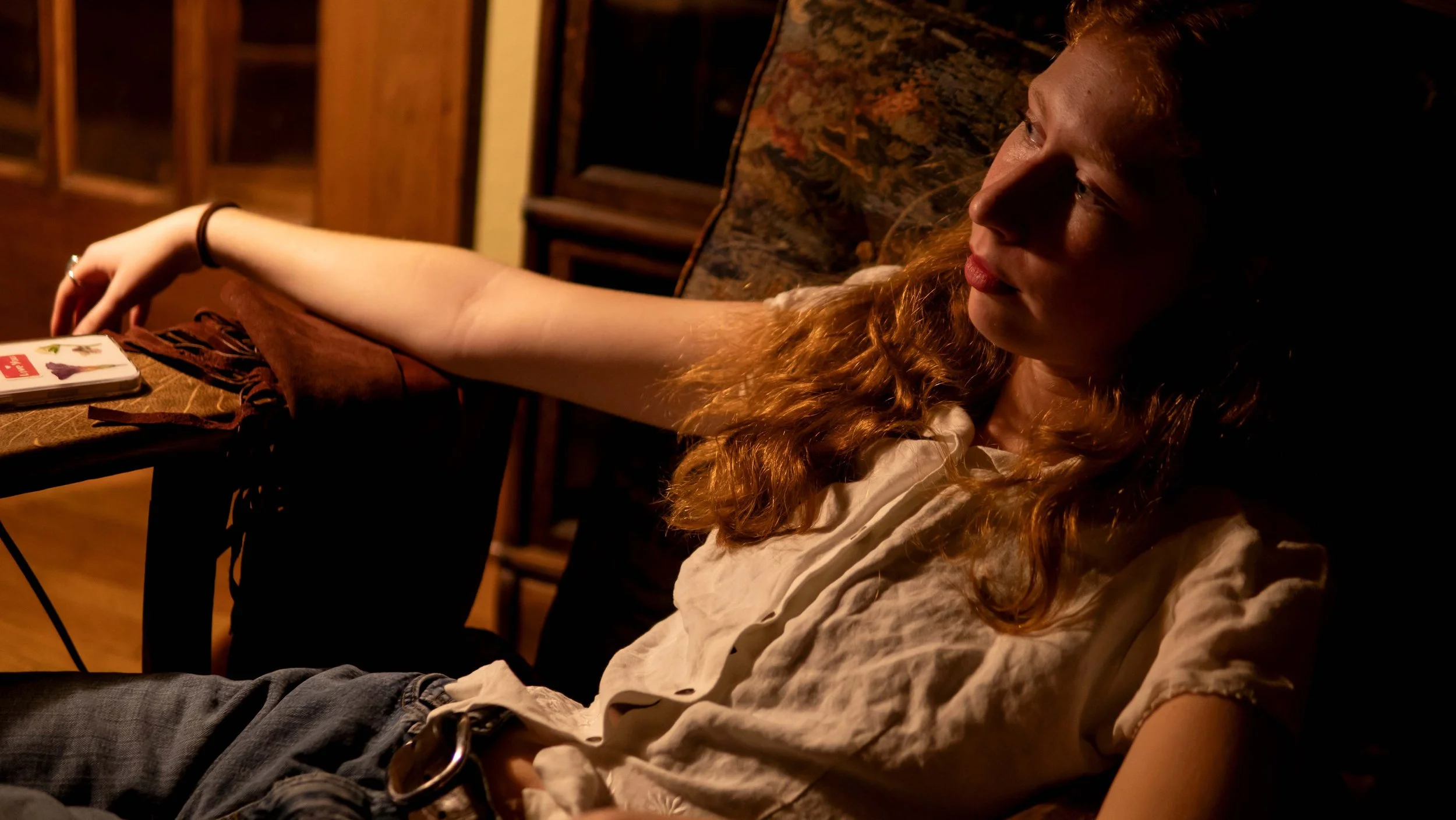 A woman with curly red hair sitting on a couch in a dimly lit room, looking to her right with a neutral expression.
