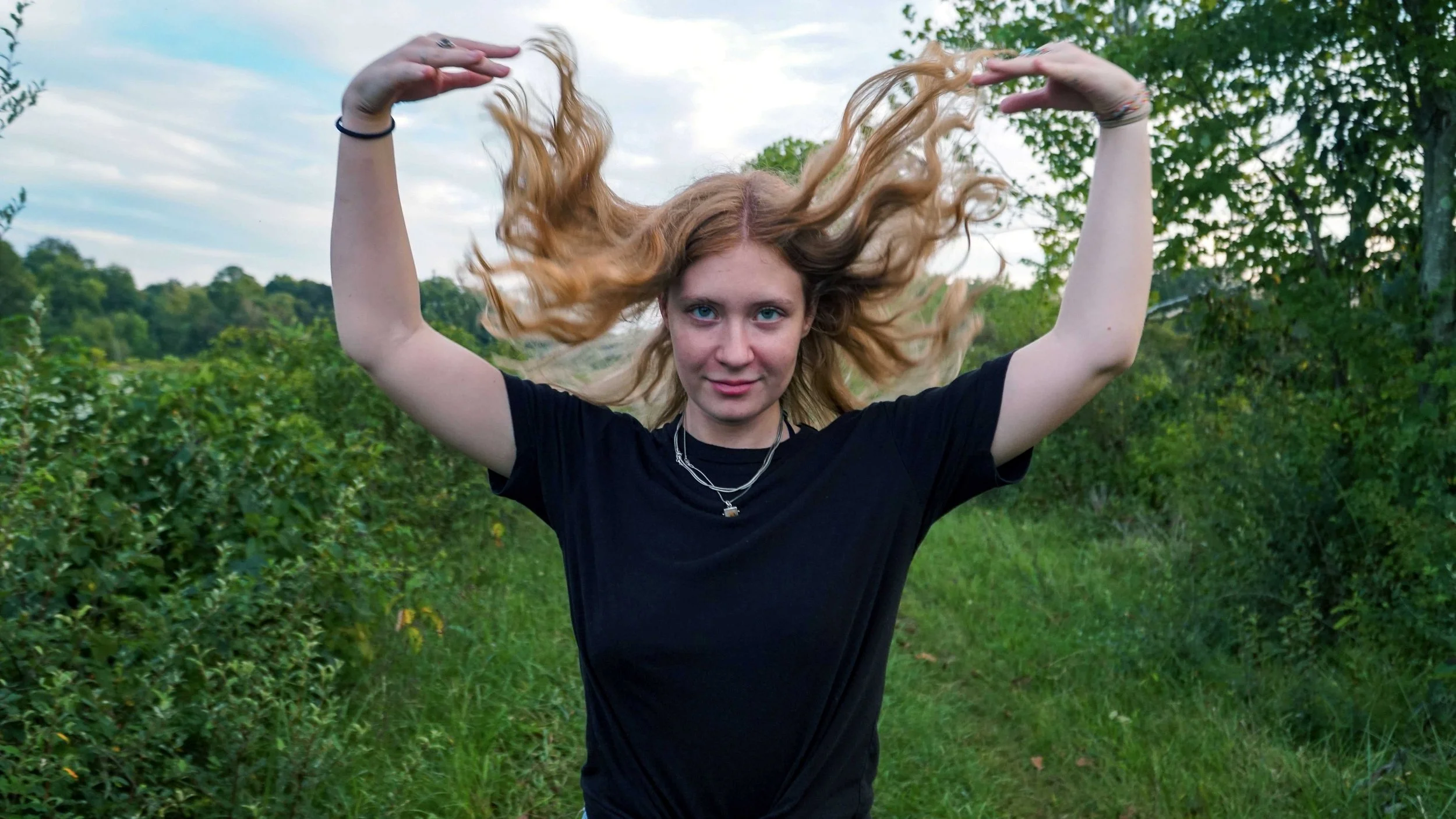 Young woman with curly red hair and blue eyes standing outdoors in a lush green area, with her arms raised and hair flying, wearing a black t-shirt and layered necklaces.