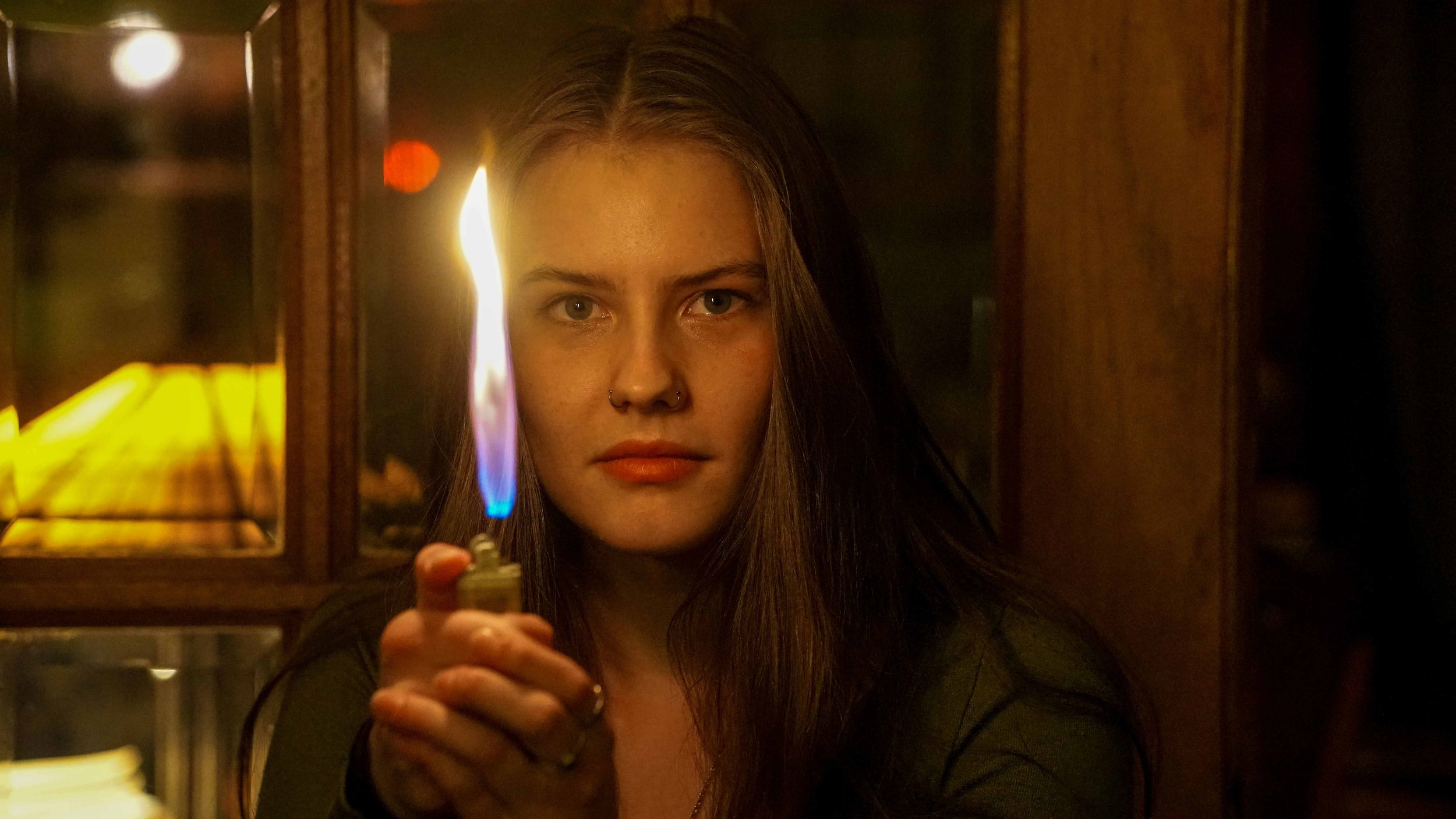 A young woman with long hair, looking directly at the camera, holds a lit lighter close to the camera with a flame. The background appears to be a dimly lit room with wooden furniture and soft lighting.