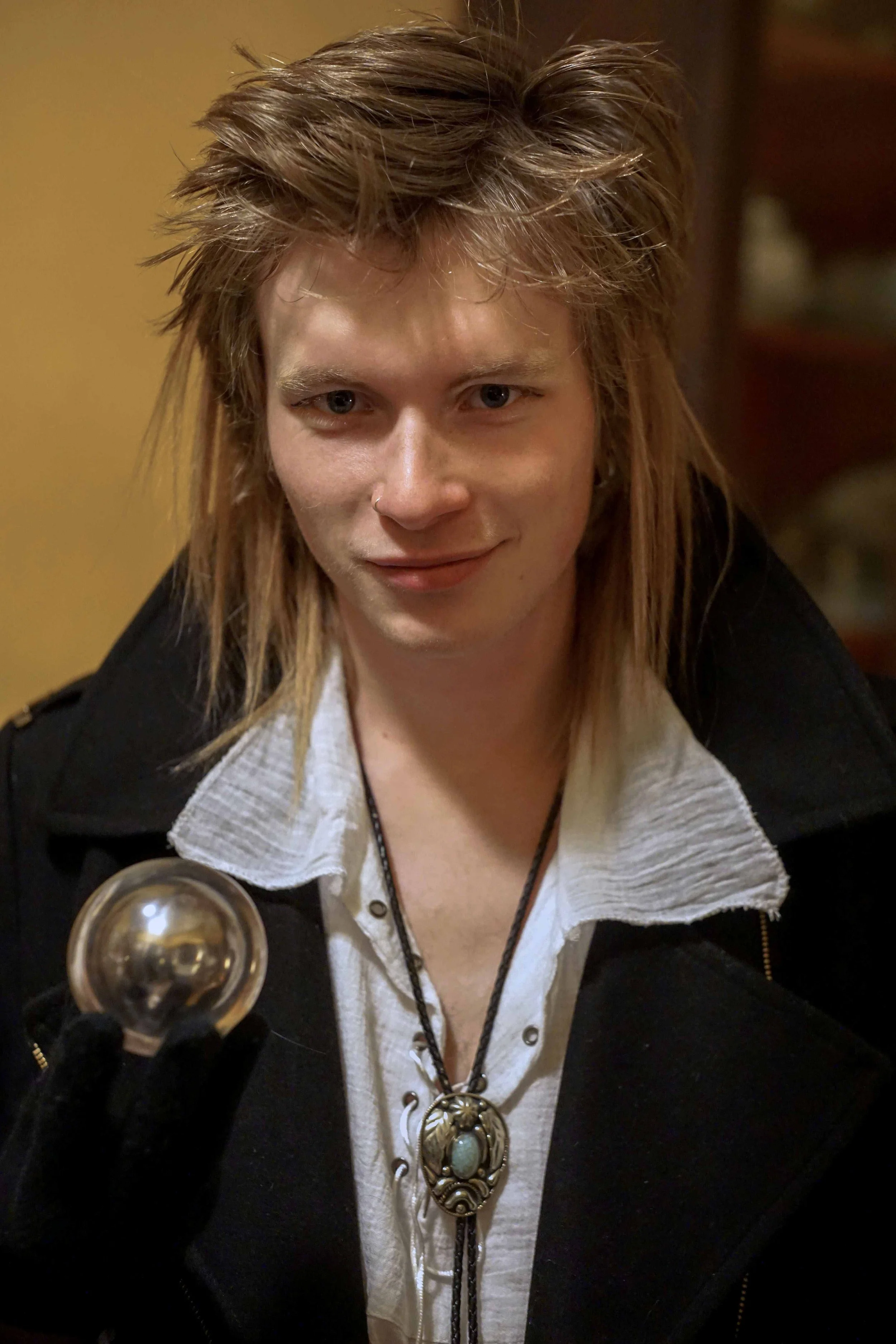 A woman with short, messy, light brown hair, wearing a black jacket and a white shirt with a ruffled collar, holding a champagne glass with a metallic lid on top.