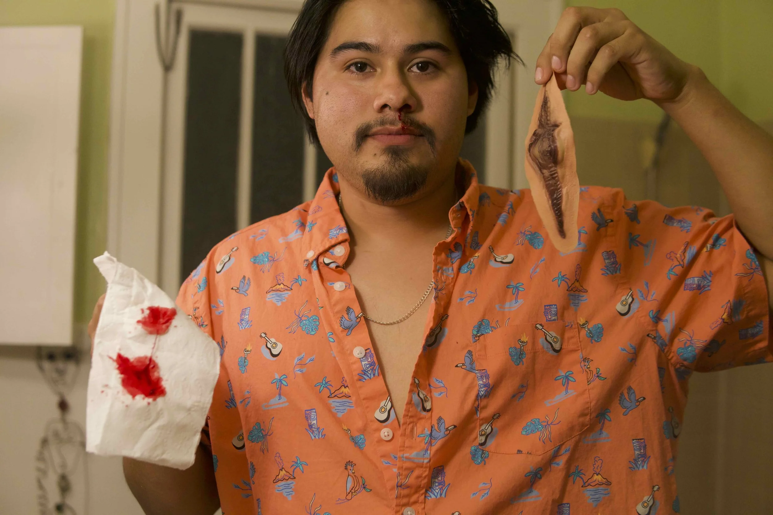 A man with dark hair, facial hair, and an orange shirt with a tropical pattern holding a piece of raw fish. The fish is cut in half, revealing its dark flesh, and has blood on the paper towel in his other hand. The background shows a kitchen setting.