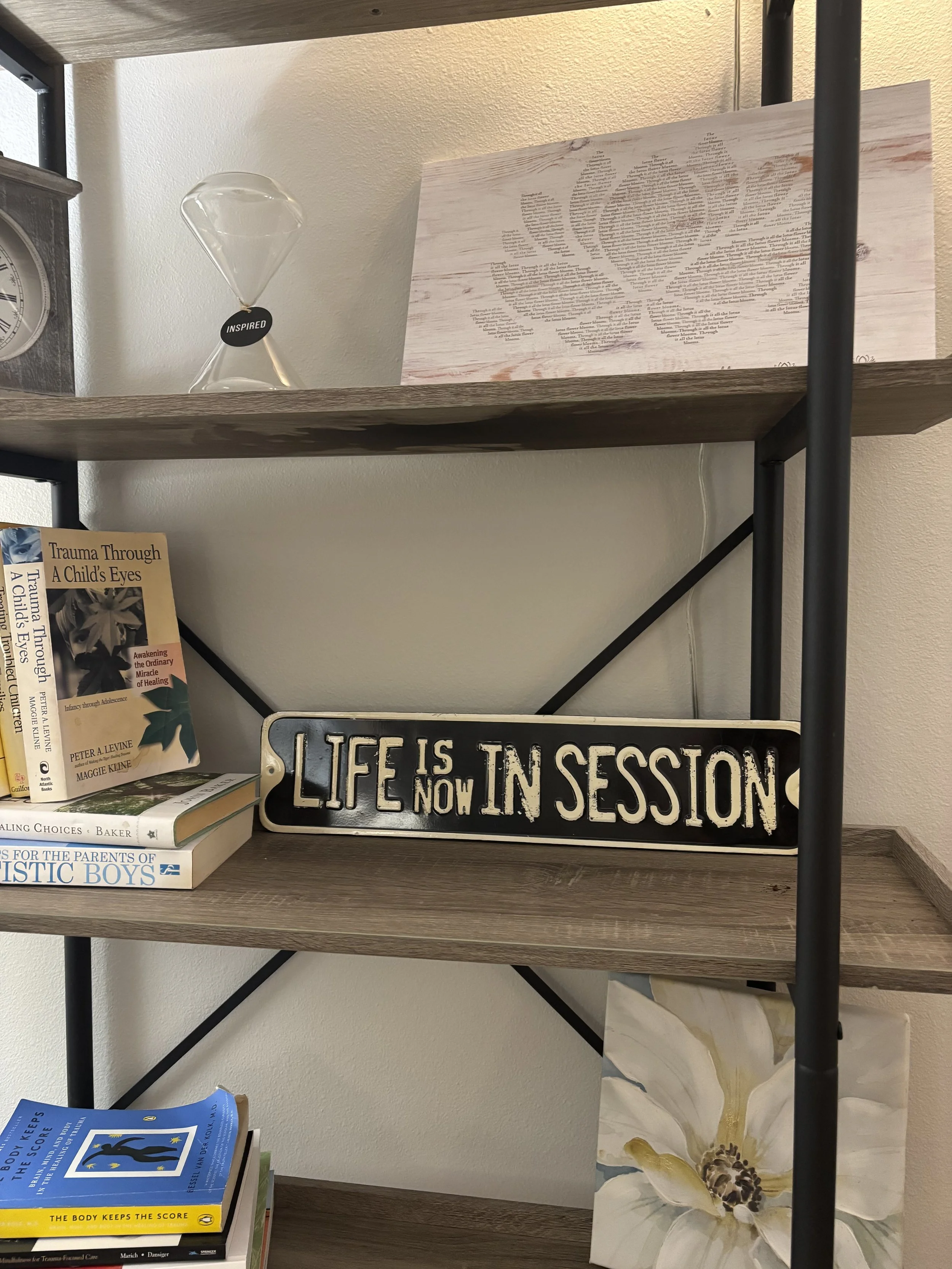Bookshelf with various books, a sign that reads 'LIFE IS IN SESSION NOW,' a floral painting, an hourglass with a tag that says 'INSPIRED,' a clock, and a wooden art piece with the word 'LOVE' composed of small text.
