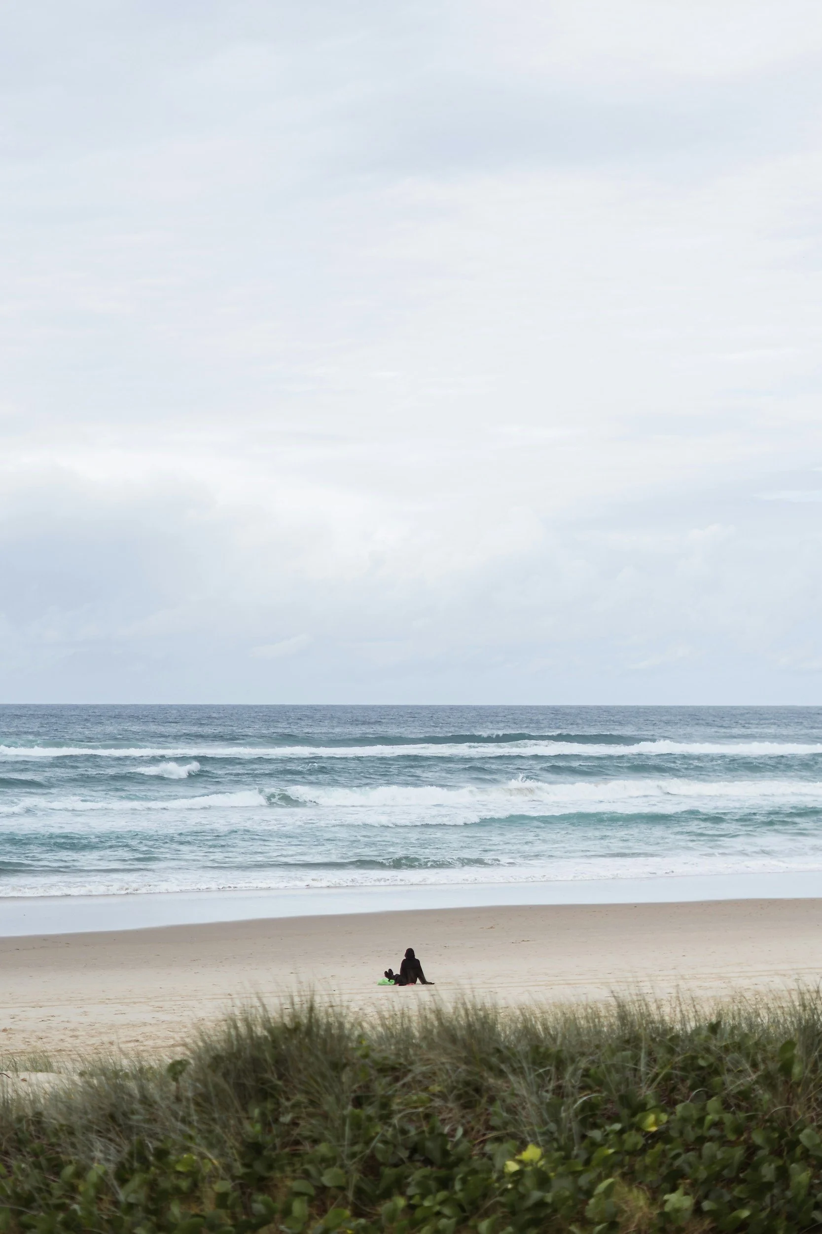 Person sitting on the beach near the water, under a cloudy sky, with green bushes in the foreground.