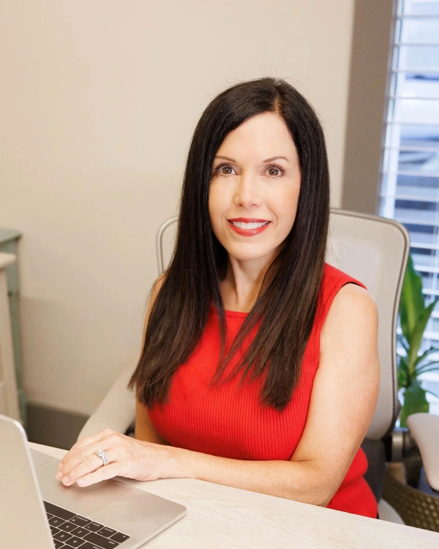 A woman with long dark hair and a red sleeveless top sitting at a desk with a silver laptop, smiling, with a window and plants in the background.