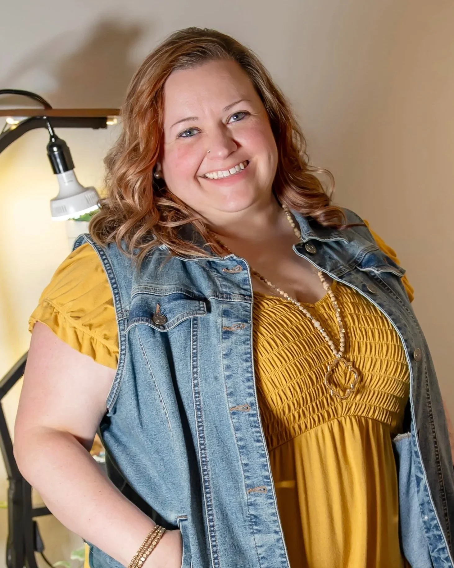 A smiling woman with red hair, wearing a yellow dress and denim vest, standing indoors near a lamp with a beige wall background.