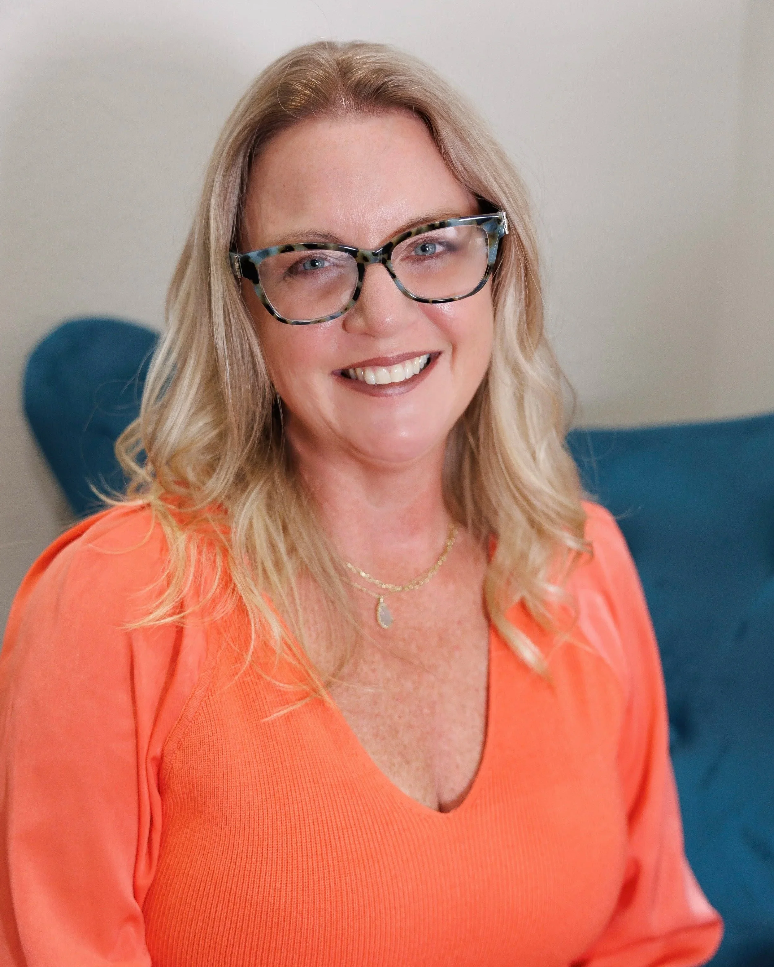 Close-up of a woman smiling, wearing glasses with a tortoiseshell pattern, and an orange top, with a blurred background.
