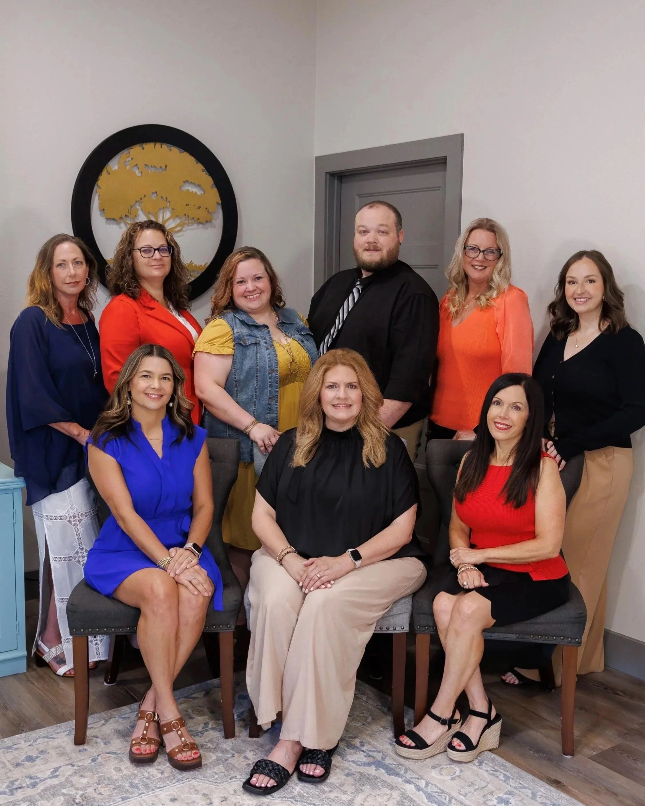A group of nine women and one man posing for a group photo in an indoor setting. The women are dressed in colorful outfits, and the man is wearing a black suit with a striped tie. They are smiling and standing or sitting around a table and chairs. There is a wall with a decorative round black and gold art piece behind them, and a gray door on the right. The floor has a patterned rug, and there are various pieces of furniture and decor in the background.