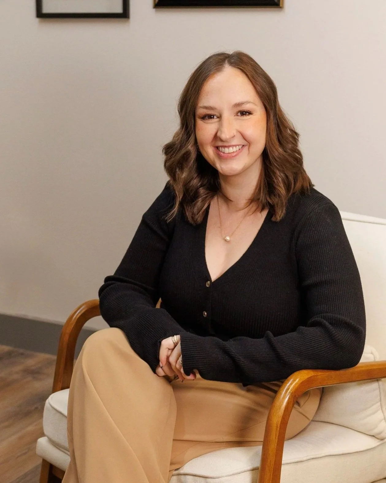 A young woman with brown hair sitting on a cream-colored armchair in a room with a white wall and framed pictures. She is smiling, wearing a black cardigan, beige pants, and a pearl necklace.