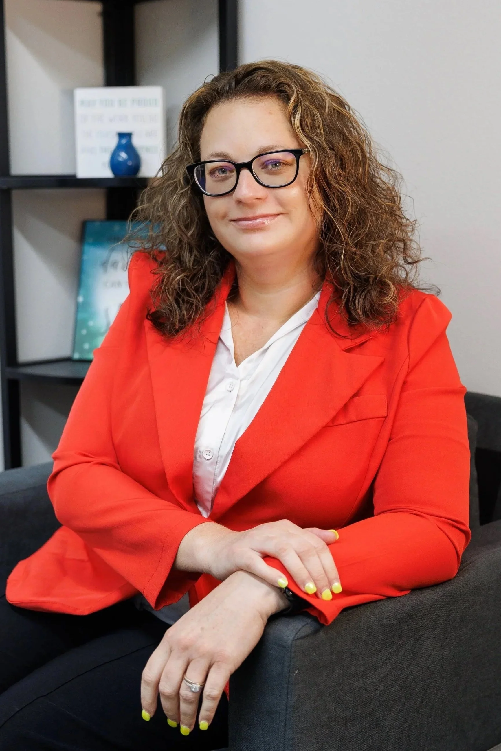 A woman with curly hair and glasses sitting at a desk. She is wearing a bright red blazer and a white shirt. The woman has a slight smile and is looking at the camera. She has yellow nail polish and a ring on her finger. There are shelves with decorative items, including a blue vase and a framed picture, in the background.