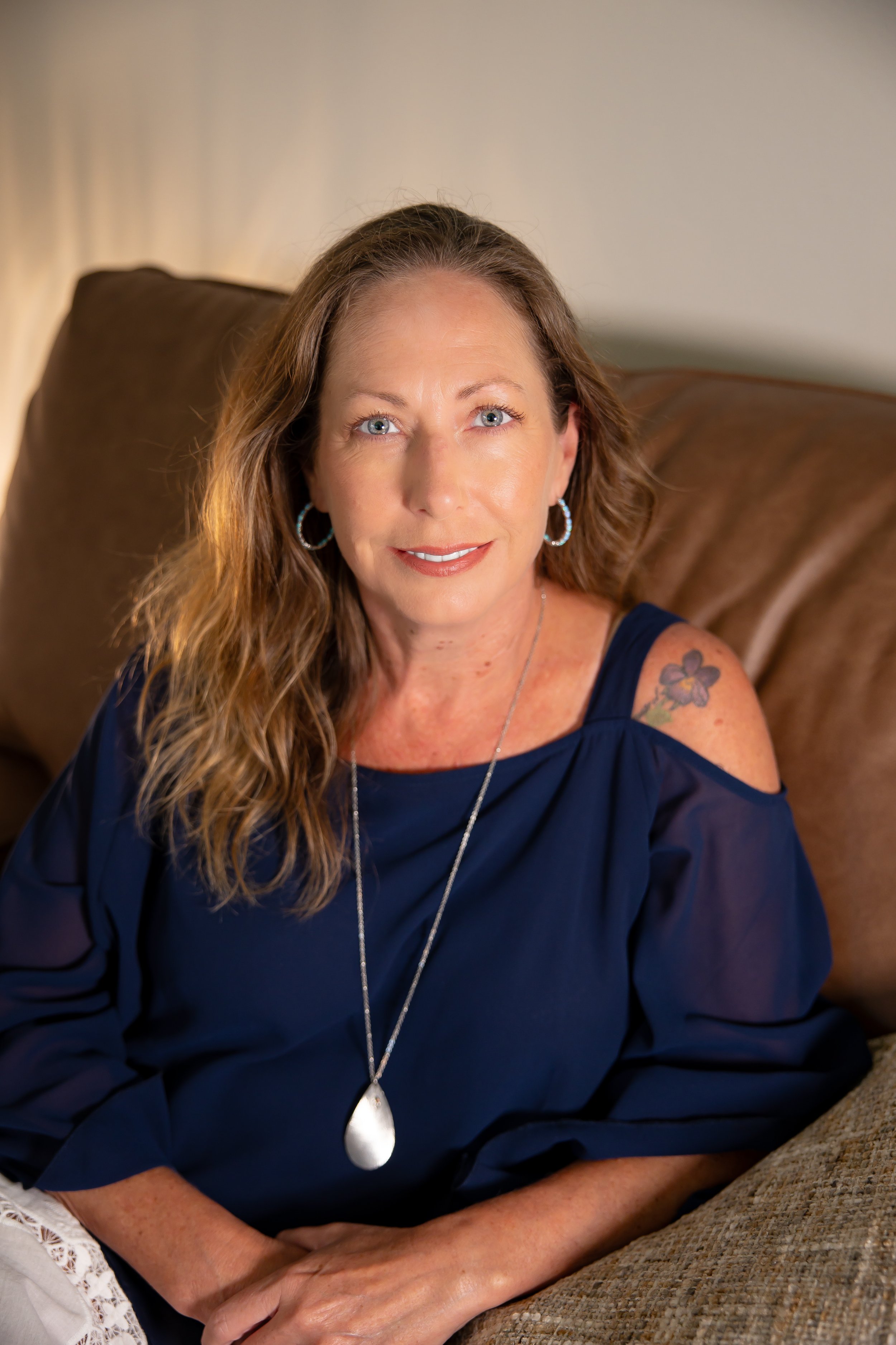 A woman with long, wavy brown hair, blue eyes, and light skin sits on a tan couch indoors. She is wearing a navy blue off-shoulder top, silver hoop earrings, and a large silver teardrop pendant necklace. She has a floral tattoo on her upper arm and is smiling softly at the camera.