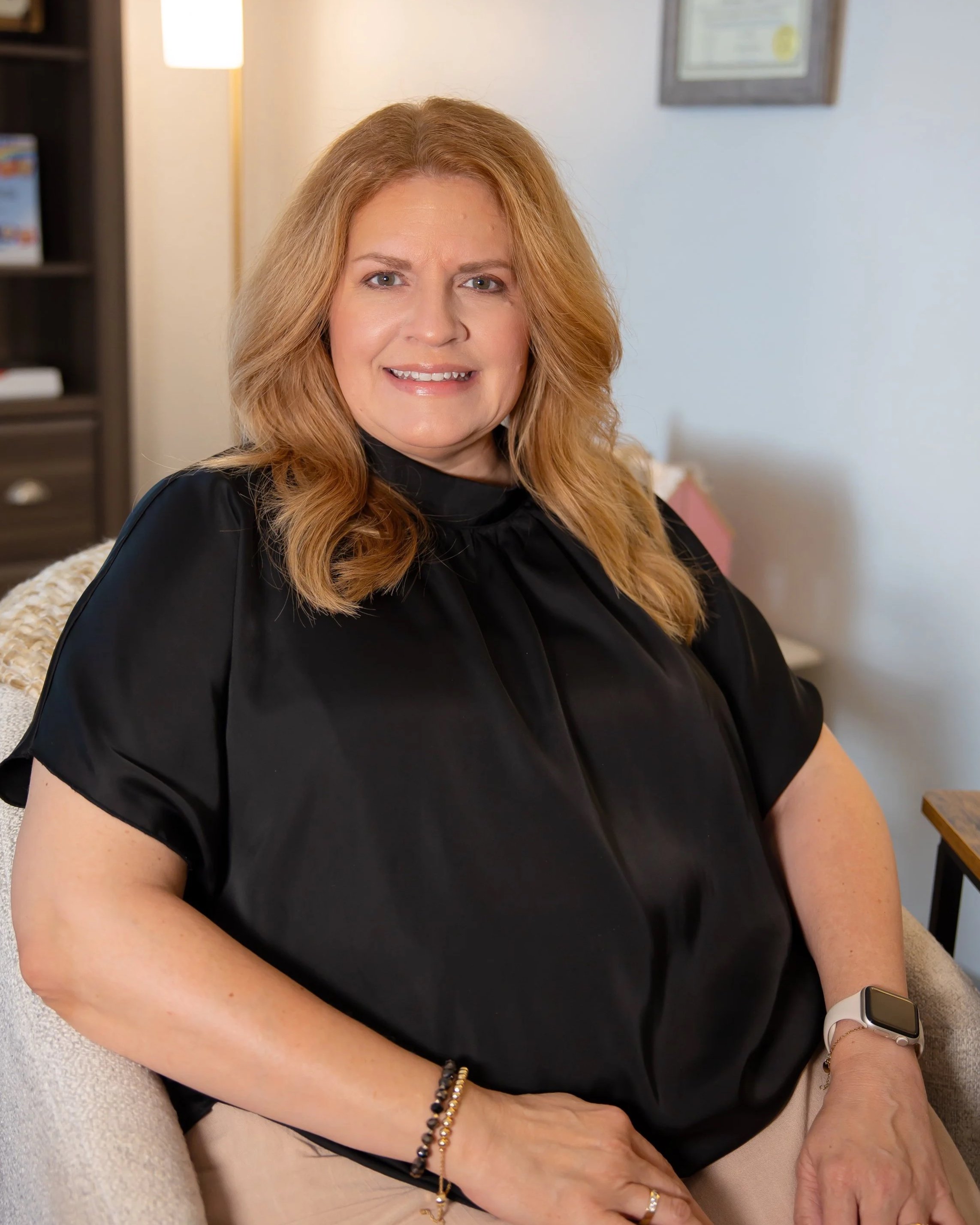 A woman with red hair and fair skin sitting on a beige couch, smiling, wearing a black top, a smartwatch, and jewelry, in a cozy room with a lamp, bookshelf, and framed pictures on the wall.