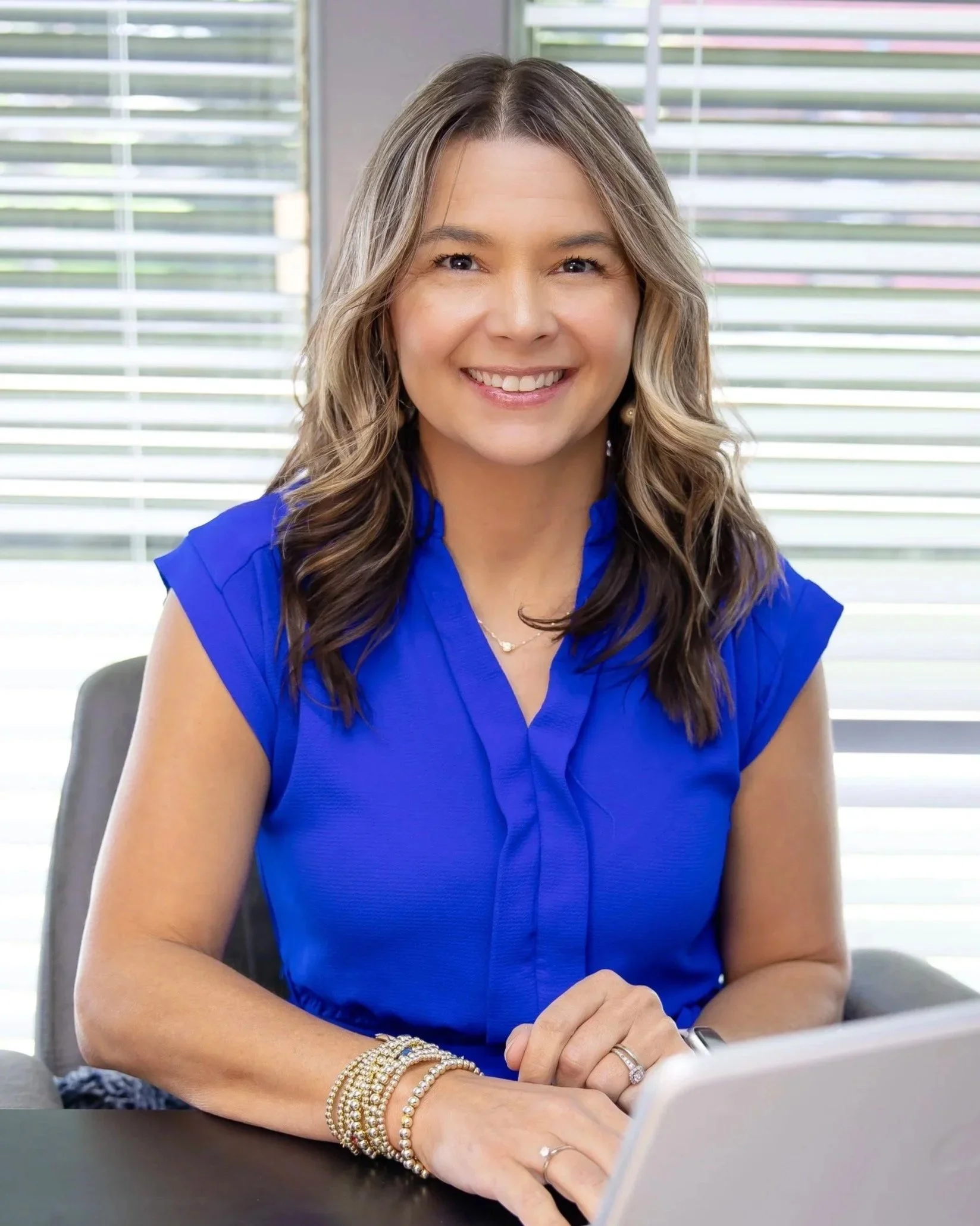 A woman with long wavy hair wearing a blue top, gold bracelets, and rings, smiling while sitting at a table with a laptop in a bright office with window blinds.