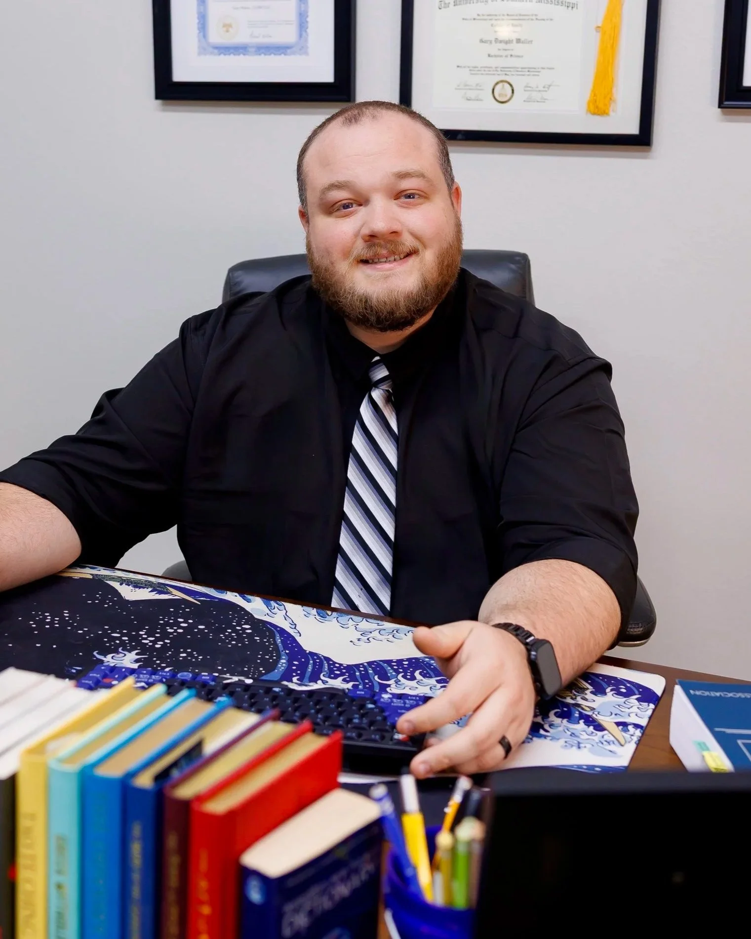 A man with a beard and short hair, wearing a black shirt and striped tie, sitting at a desk with framed certificates on the wall behind him. There are colorful books and a game or puzzle on the desk in front of him.