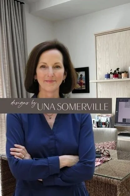 A woman with shoulder-length brown hair, wearing a blue shirt, standing in a room with a desk, computer, shelves with plants and decor, and a framed photograph on the wall.
