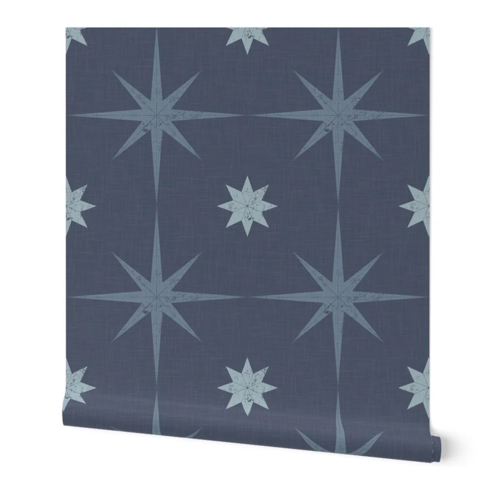 Denim-blue textured wallpaper roll featuring a repeating pattern of large, distressed eight-pointed compass stars and starbursts in pale ice blue.