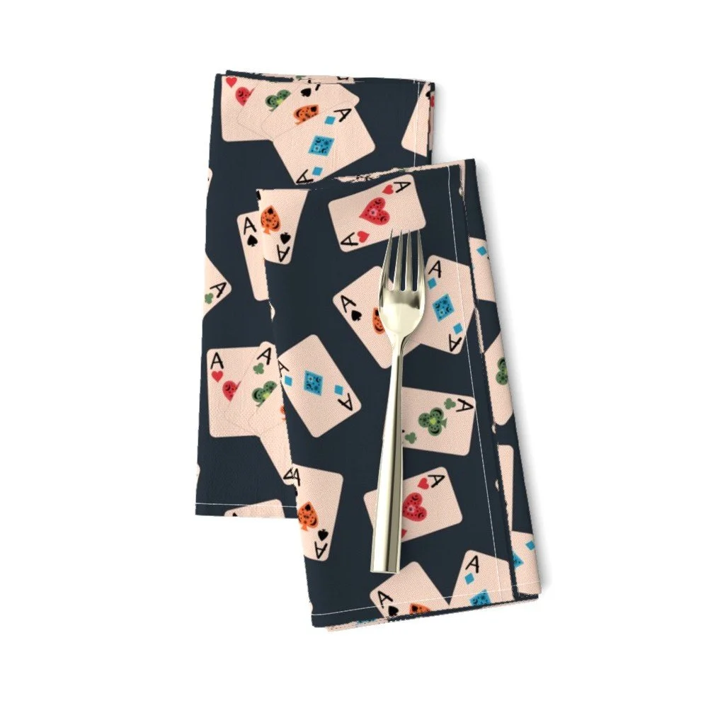 Folded dark gray cloth dinner napkins with a silver fork on top, patterned with scattered playing cards displaying whimsical aces of all four suits.