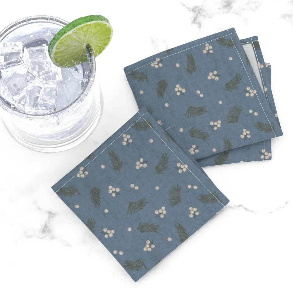 A glass of water with ice cubes and a lime wedge on the rim, placed on a white surface next to three blue napkins with a white floral and green leaf pattern.