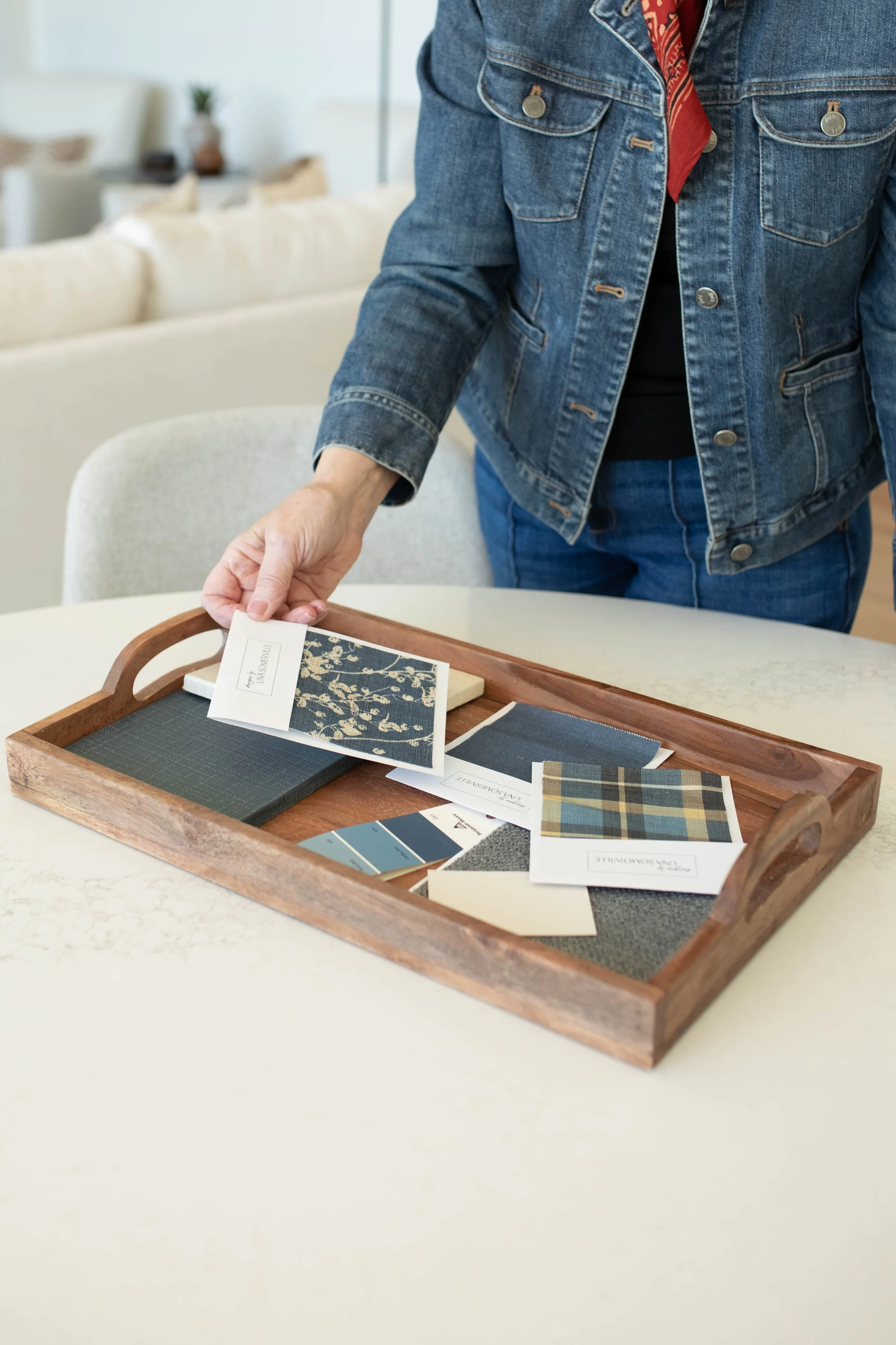 Person wearing a denim jacket and black shirt standing over a wooden tray with fabric and color samples on a white table.