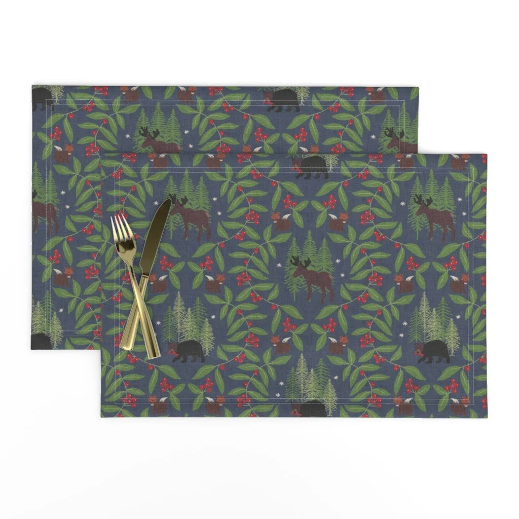 Alt text: A table place setting with gold cutlery on navy-blue placemats printed with woodland wildlife including moose, black bears, foxes, pine trees, and red winter berries.