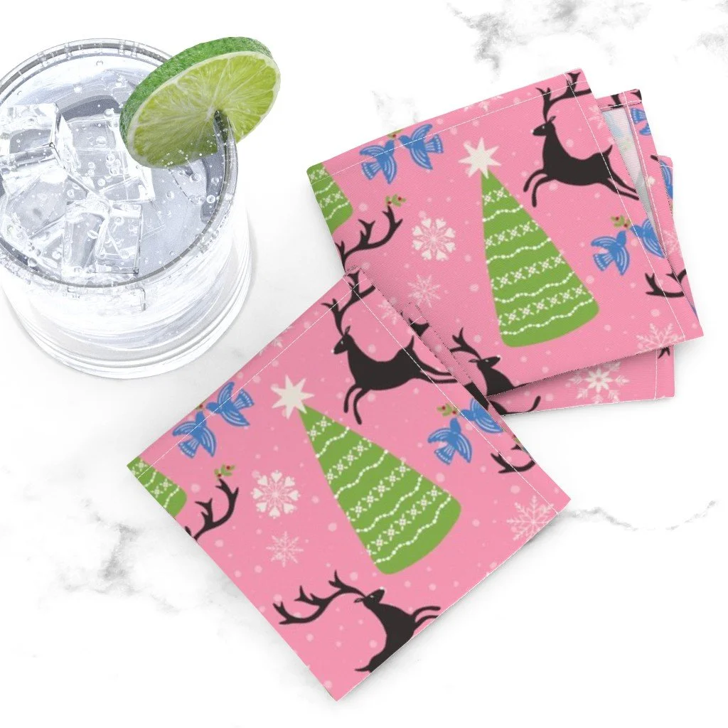 An iced drink with a lime wheel on white marble beside pink cloth cocktail napkins printed with leaping black reindeer, green Christmas trees, and bluebirds.