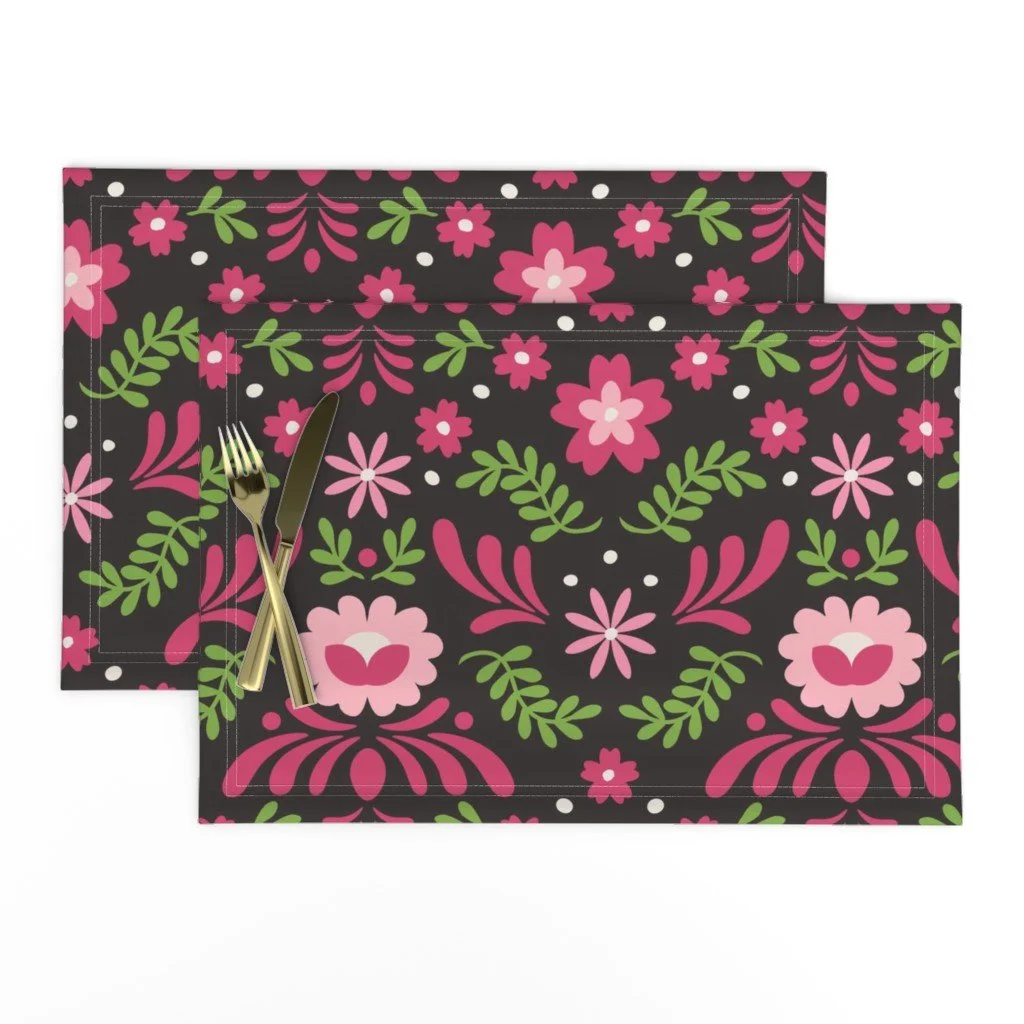 A place setting with gold cutlery on dark chocolate  brown placemats printed with vibrant pink and green Scandinavian-style folk art floral motifs.