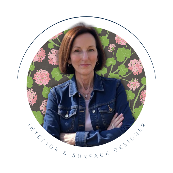 Una Somerville  - interior and surface designer in a jean jacket and pink geranium textile pattern in the background.ngth brown hair, wearing a denim jacket and crossing her arms, poses in front of a patterned background.