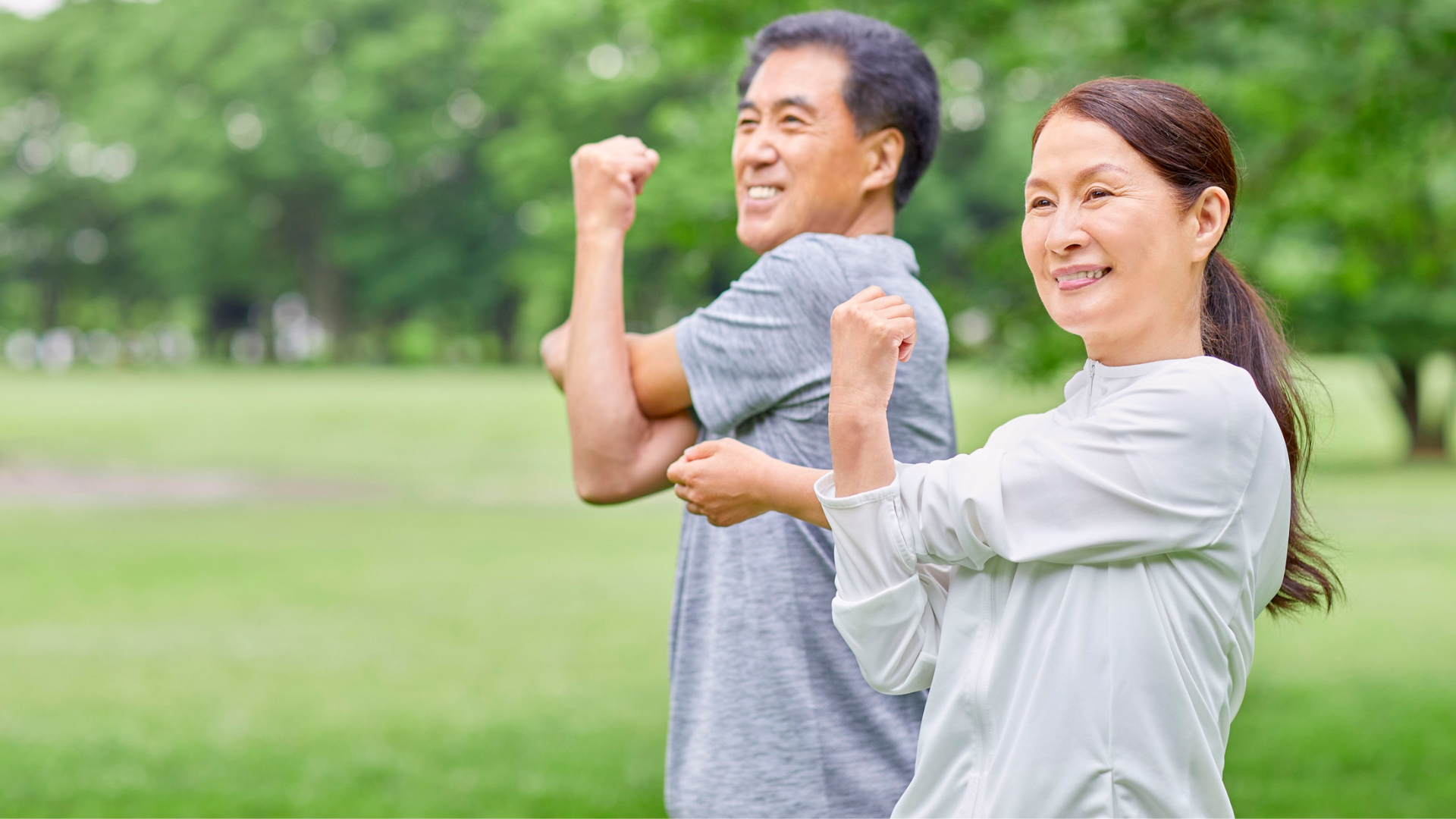 Smiling man and woman outdoors, flexing muscles in a park with green trees.