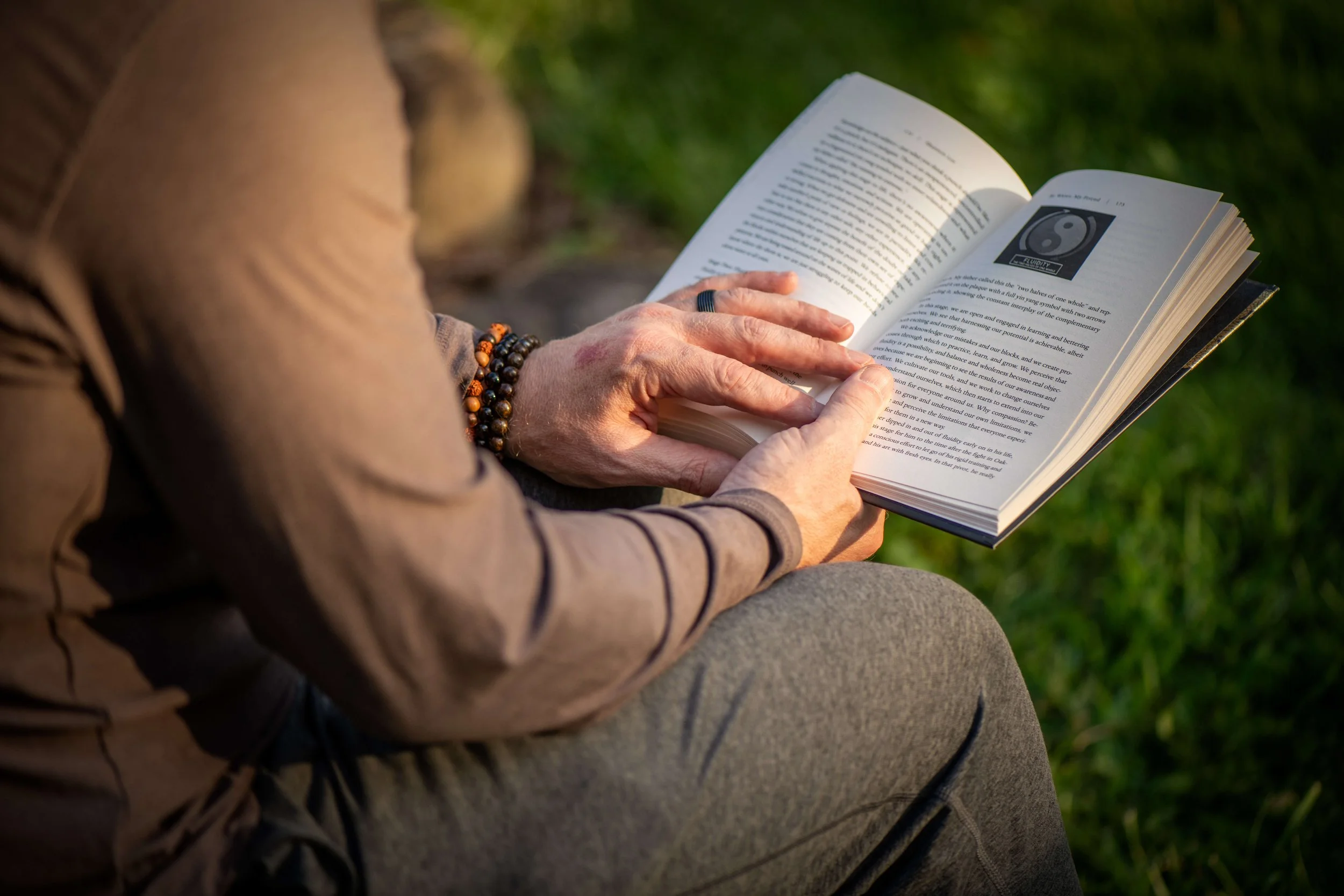 A person sitting outdoors, reading a book with a yin-yang symbol on one of its pages.