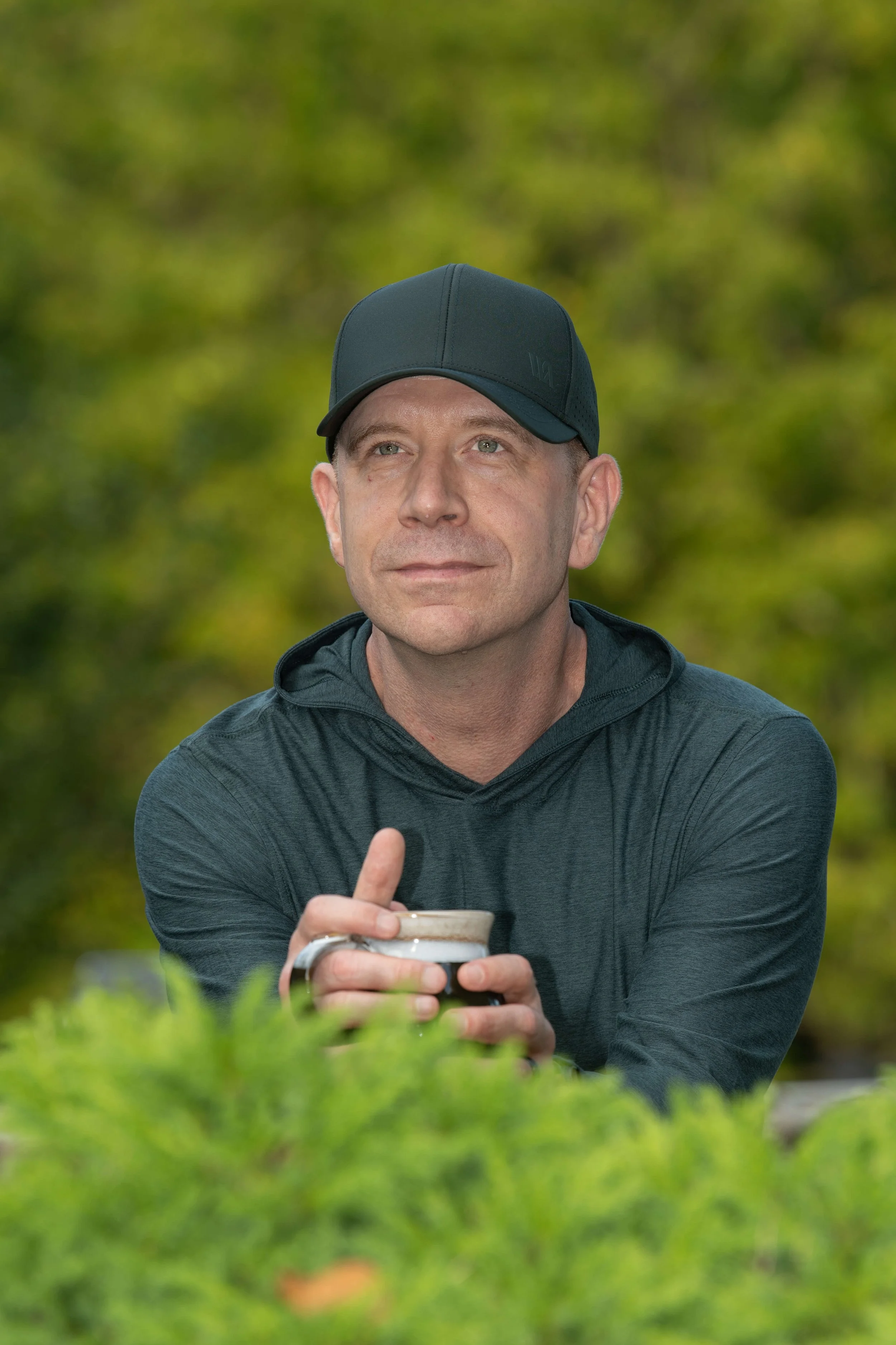 A man wearing a dark baseball cap and jacket, holding a coffee cup, sitting outdoors surrounded by green foliage, gazing upward.