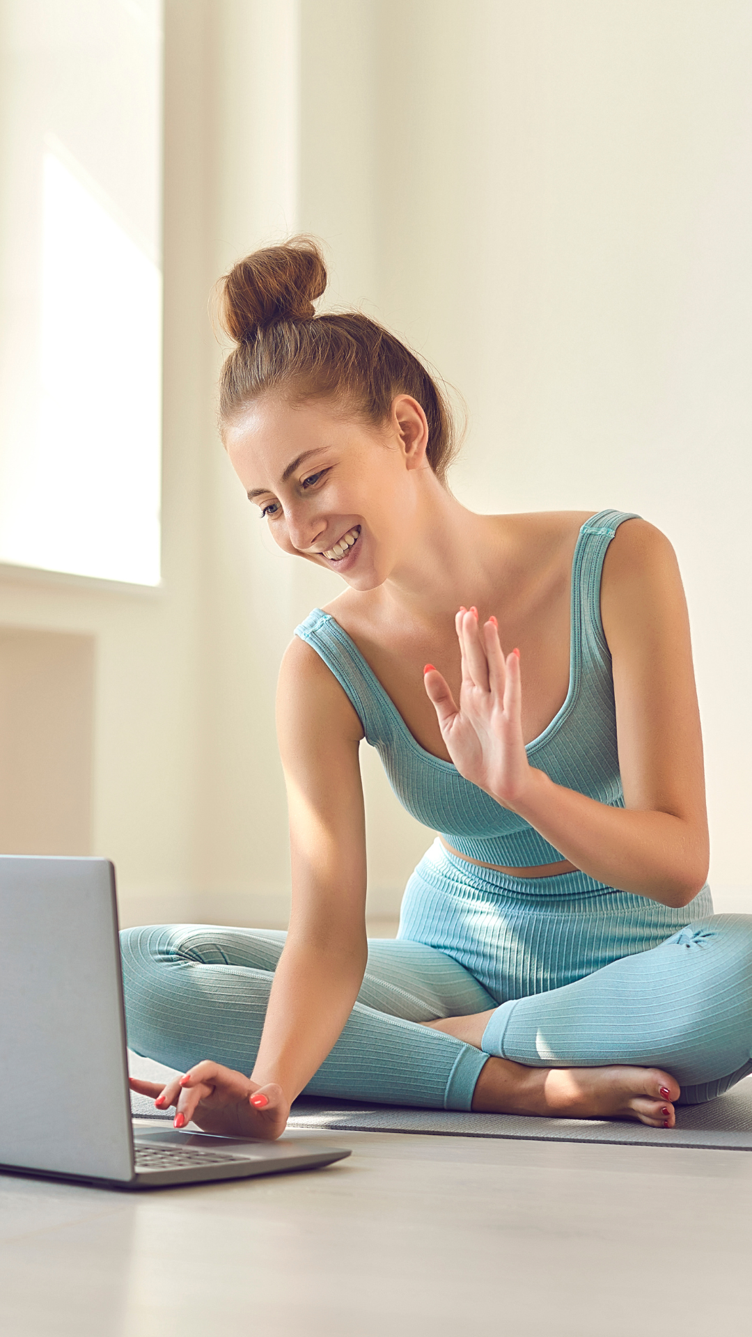 Young woman practicing yoga at home, sitting cross-legged on the floor, waving at a laptop