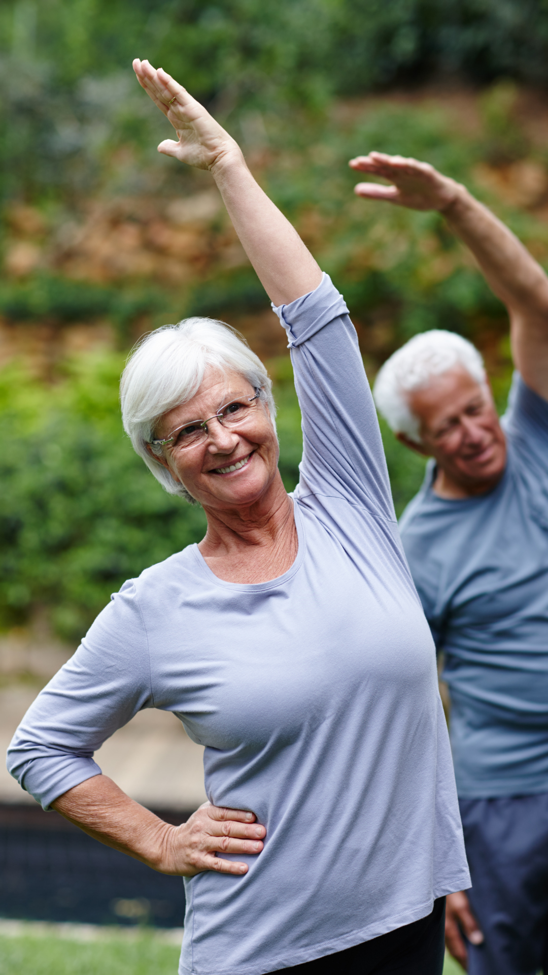 An elderly woman wearing glasses and a gray long-sleeve shirt is smiling and stretching outdoors. There is a man behind her, also stretching, in a similar gray shirt. The background is blurred with green trees and bushes.
