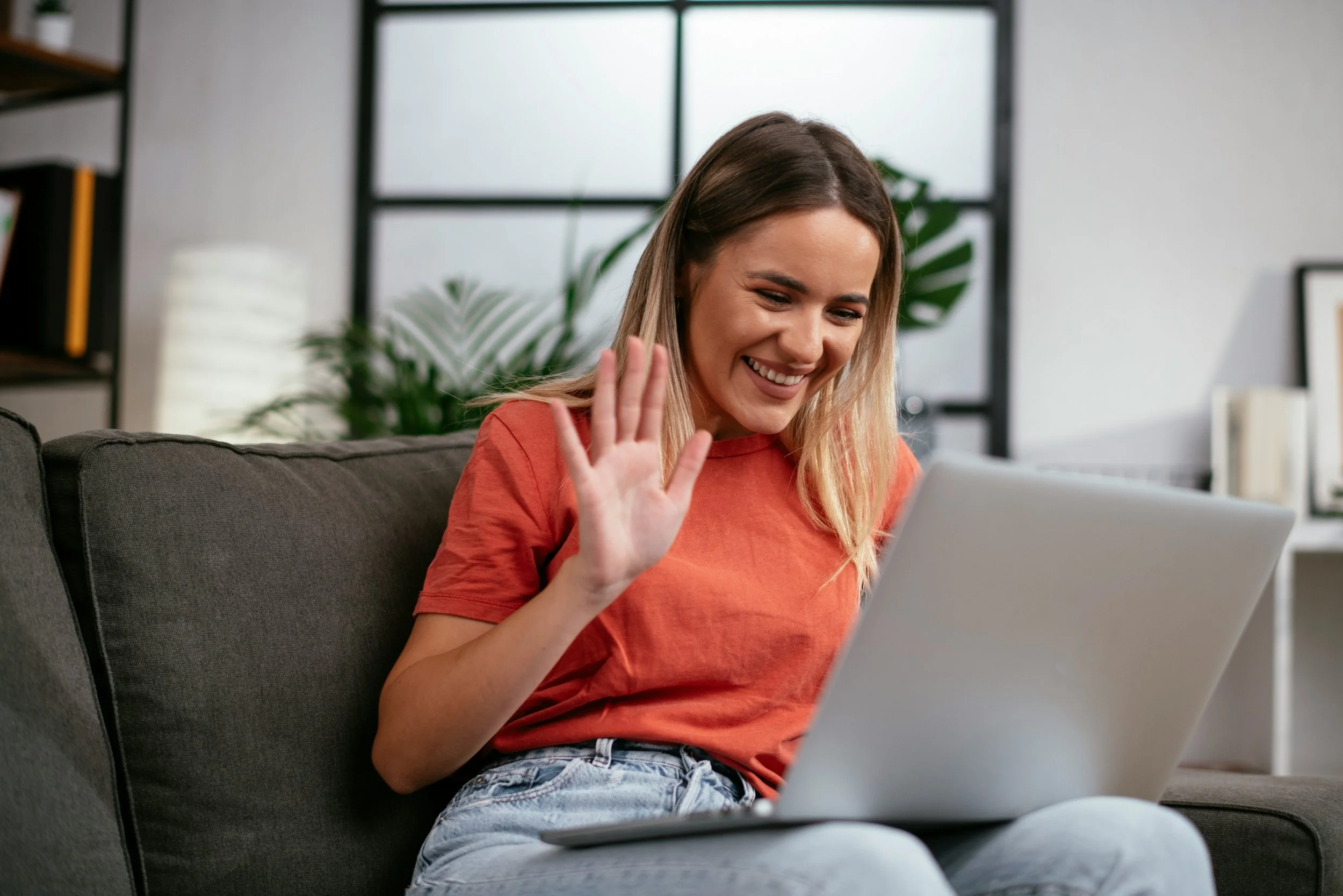 A young woman with long blonde hair wearing an orange t-shirt and light blue jeans sitting on a gray couch, smiling, and waving at a laptop in front of her in a modern room with large windows and plants.