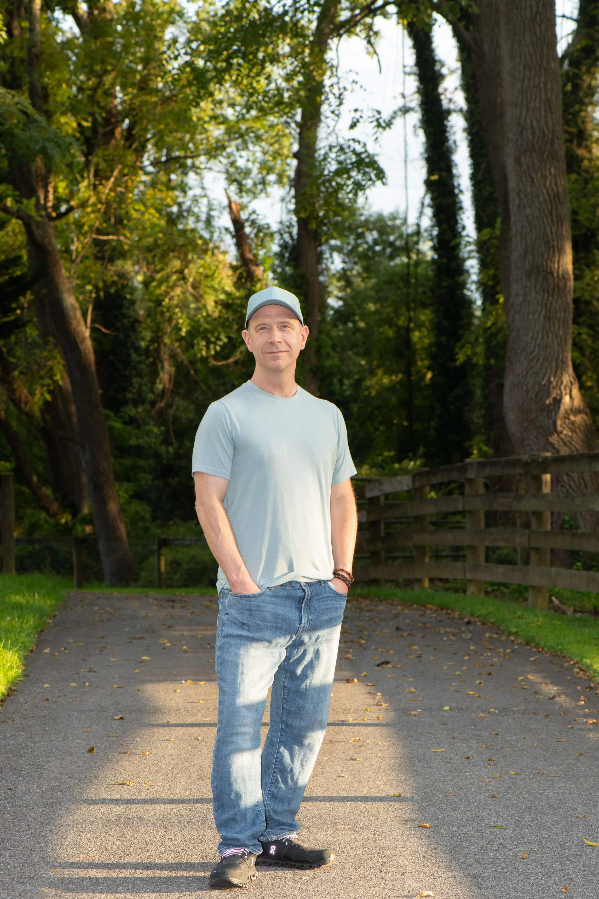 A man standing outdoors on a paved path during the daytime, with trees and greenery in the background, wearing a light blue cap, light gray t-shirt, ripped jeans, and black athletic shoes. He has his hands in his pockets and is smiling slightly.