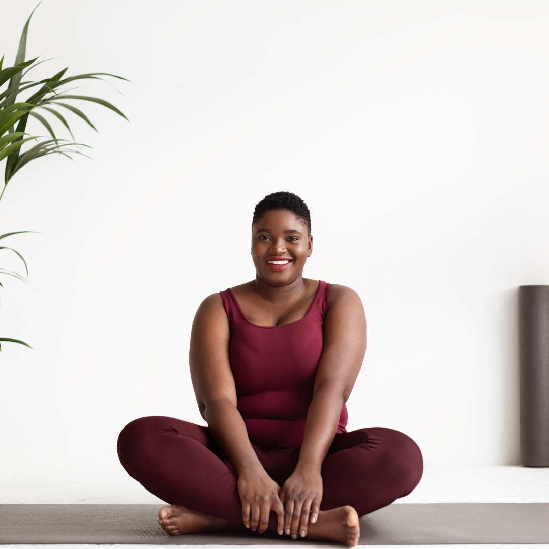 A woman sitting cross-legged on a yoga mat, smiling, in a minimalist room with a white wall and a green plant.
