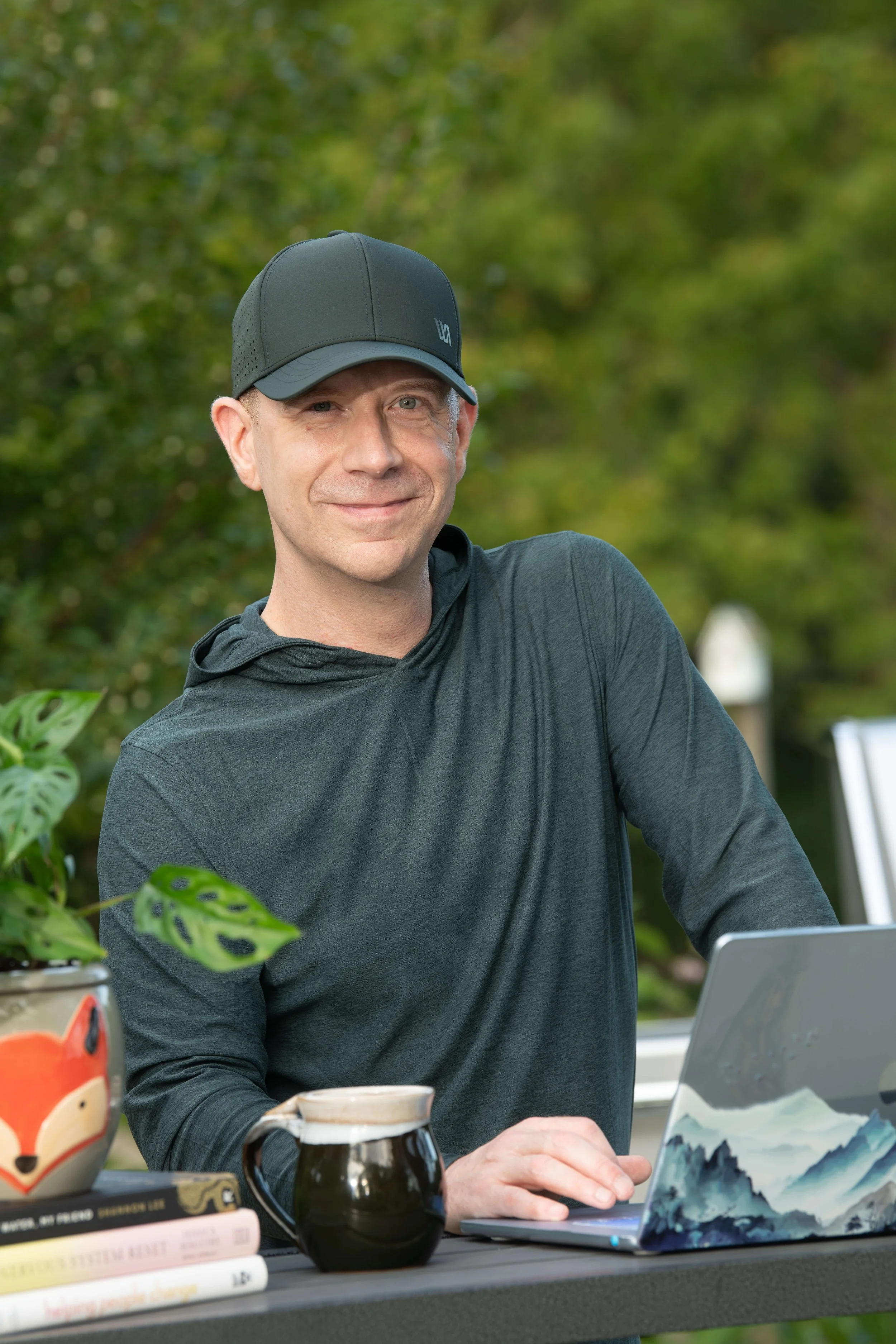 A man in a black baseball cap and dark hoodie sitting at an outdoor table with a laptop, mugs, and books, surrounded by greenery.