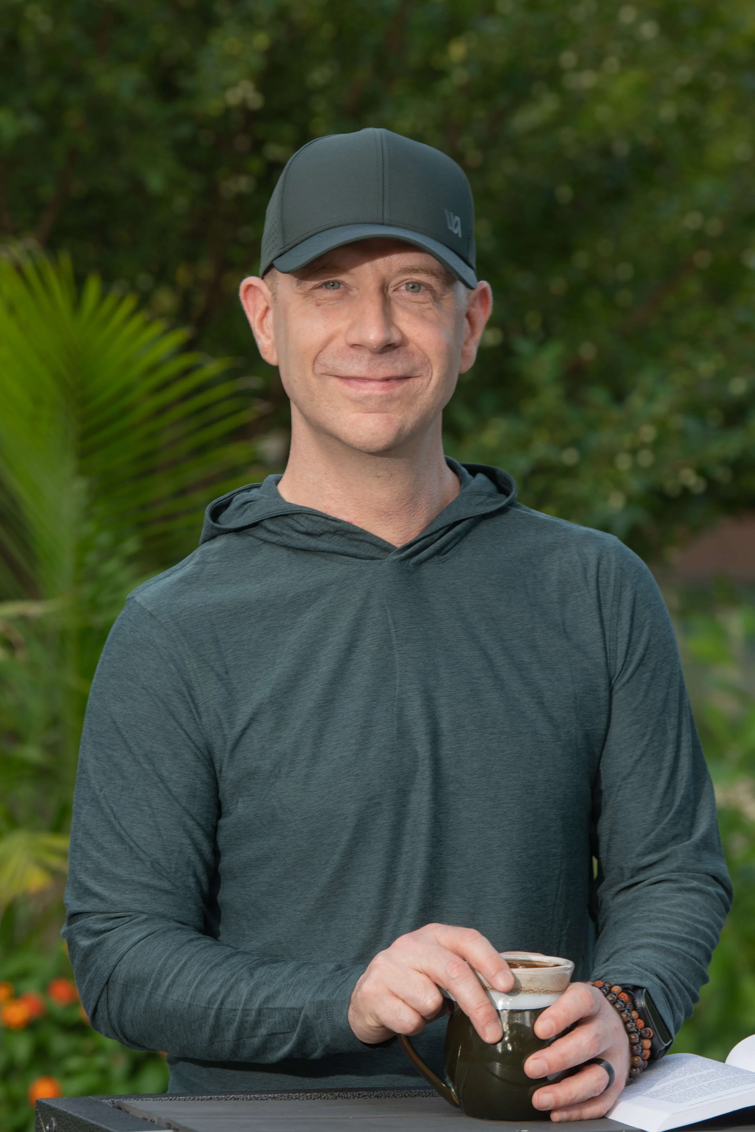 A man in a gray cap and gray hoodie holding a coffee pot and a cup, standing outdoors with green trees in the background.
