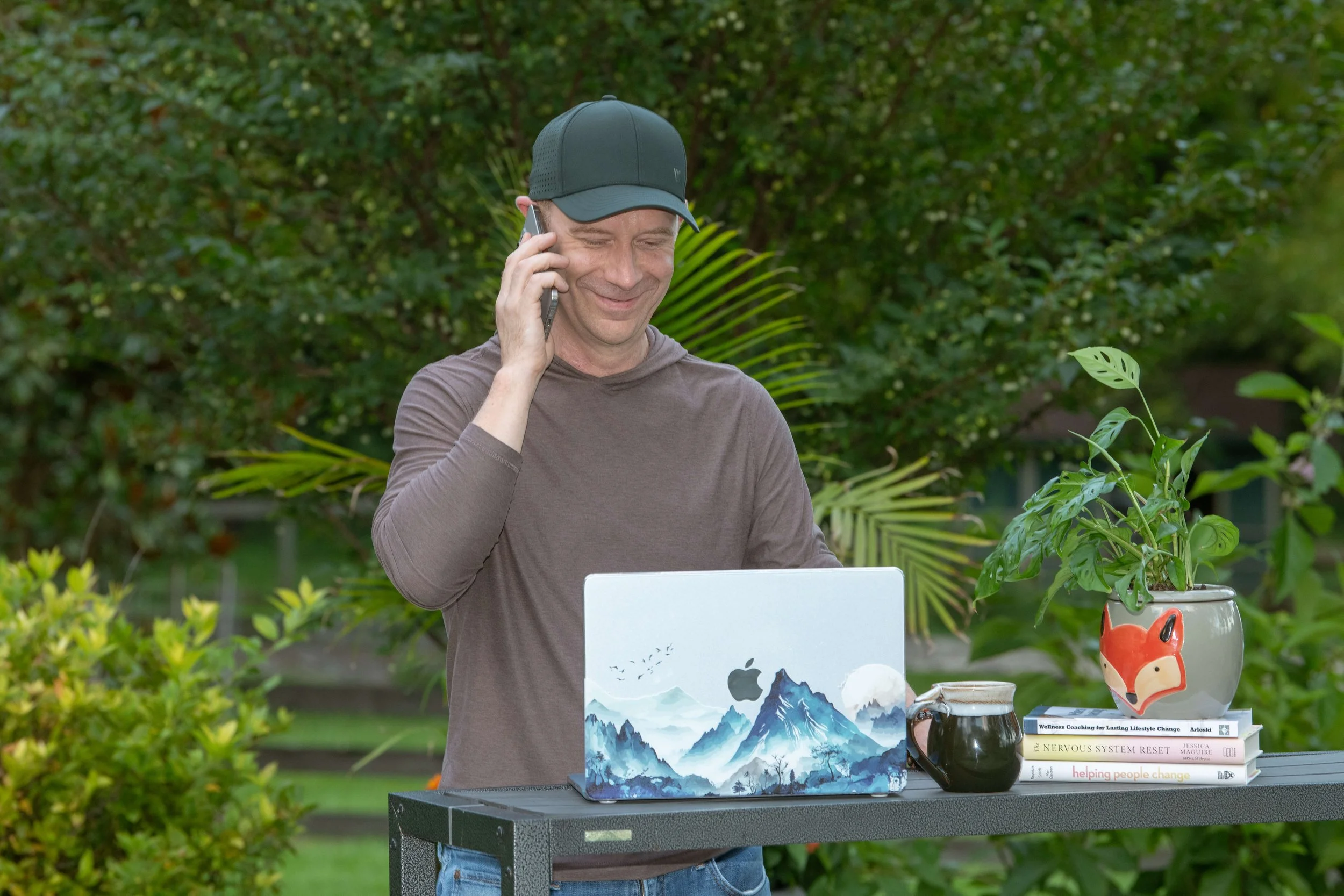Man in a black cap smiling while talking on a phone, standing outdoors with greenery in background, next to a table with books, a painting of mountains, a pig-shaped vase, and a mug.