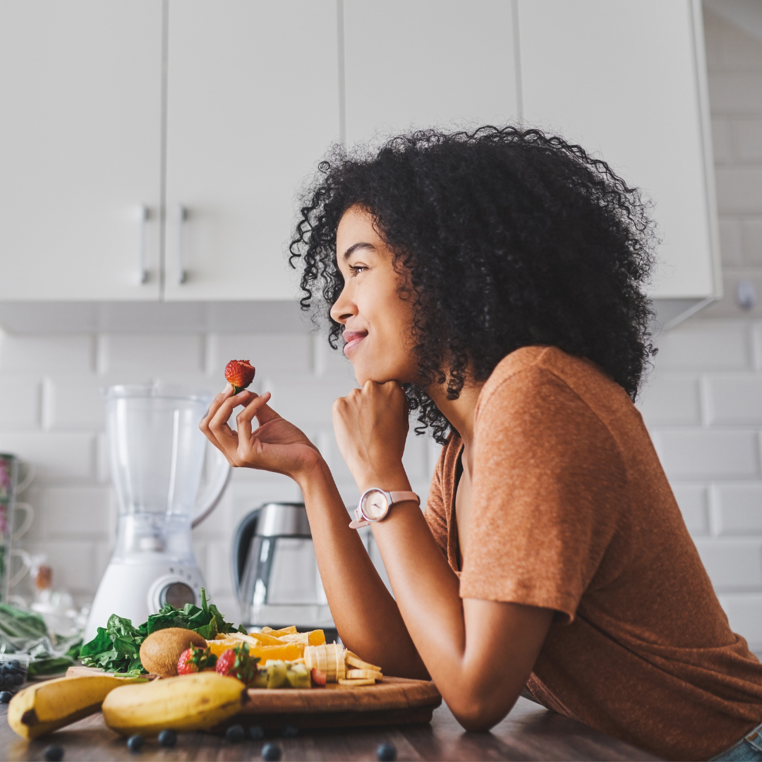 A woman with curly hair and a watch is holding a strawberry and smiling in a kitchen next to a cutting board with bananas, strawberries, kiwi, and leafy greens.