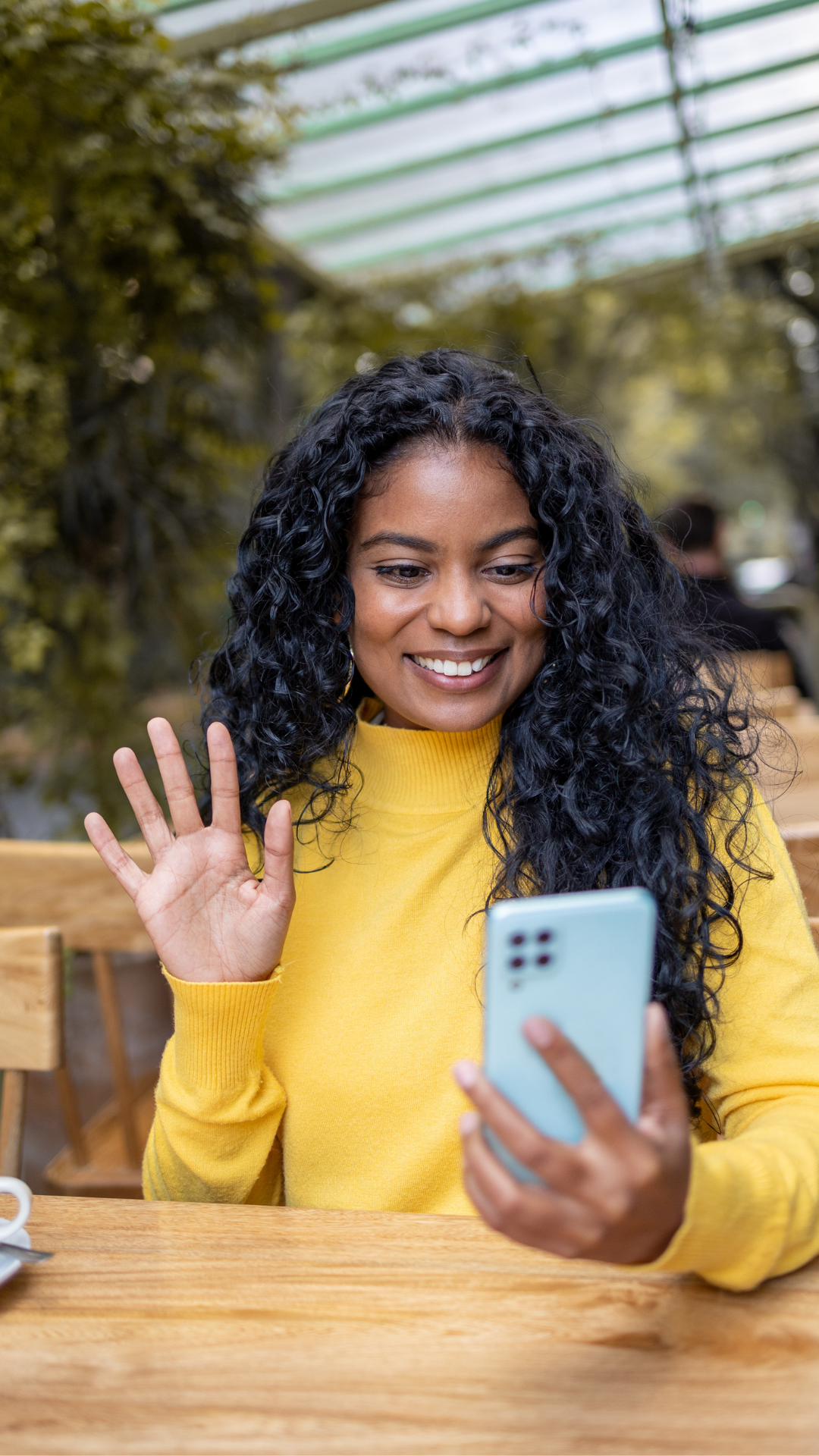 A woman in a yellow sweater with curly black hair holding a smartphone and waving while sitting at a wooden table in a greenhouse or botanical garden.