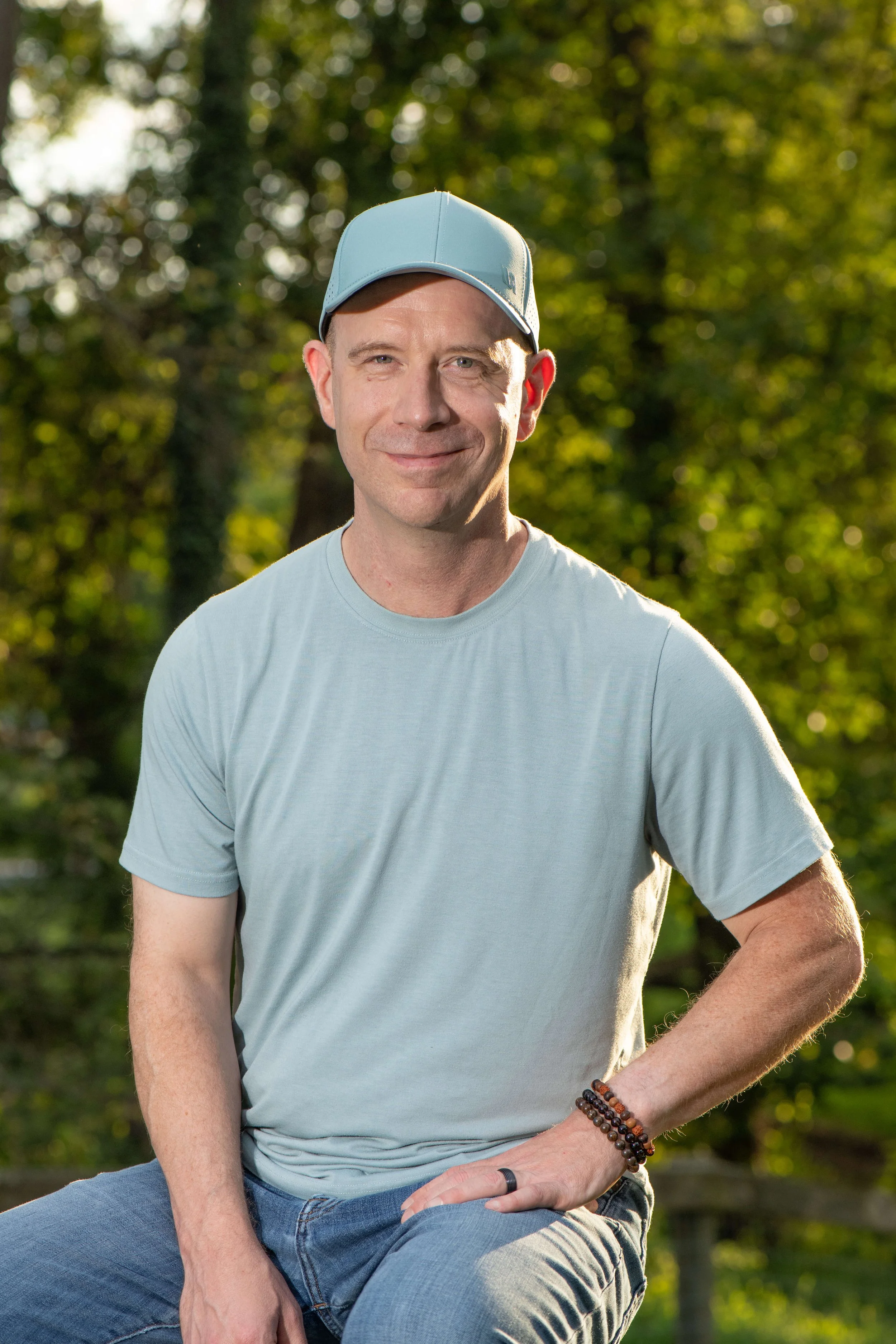 A man sitting outdoors in a park, wearing a light blue baseball cap, a light gray t-shirt, and multiple beaded bracelets on his right wrist, smiling at the camera with a background of out-of-focus green trees.